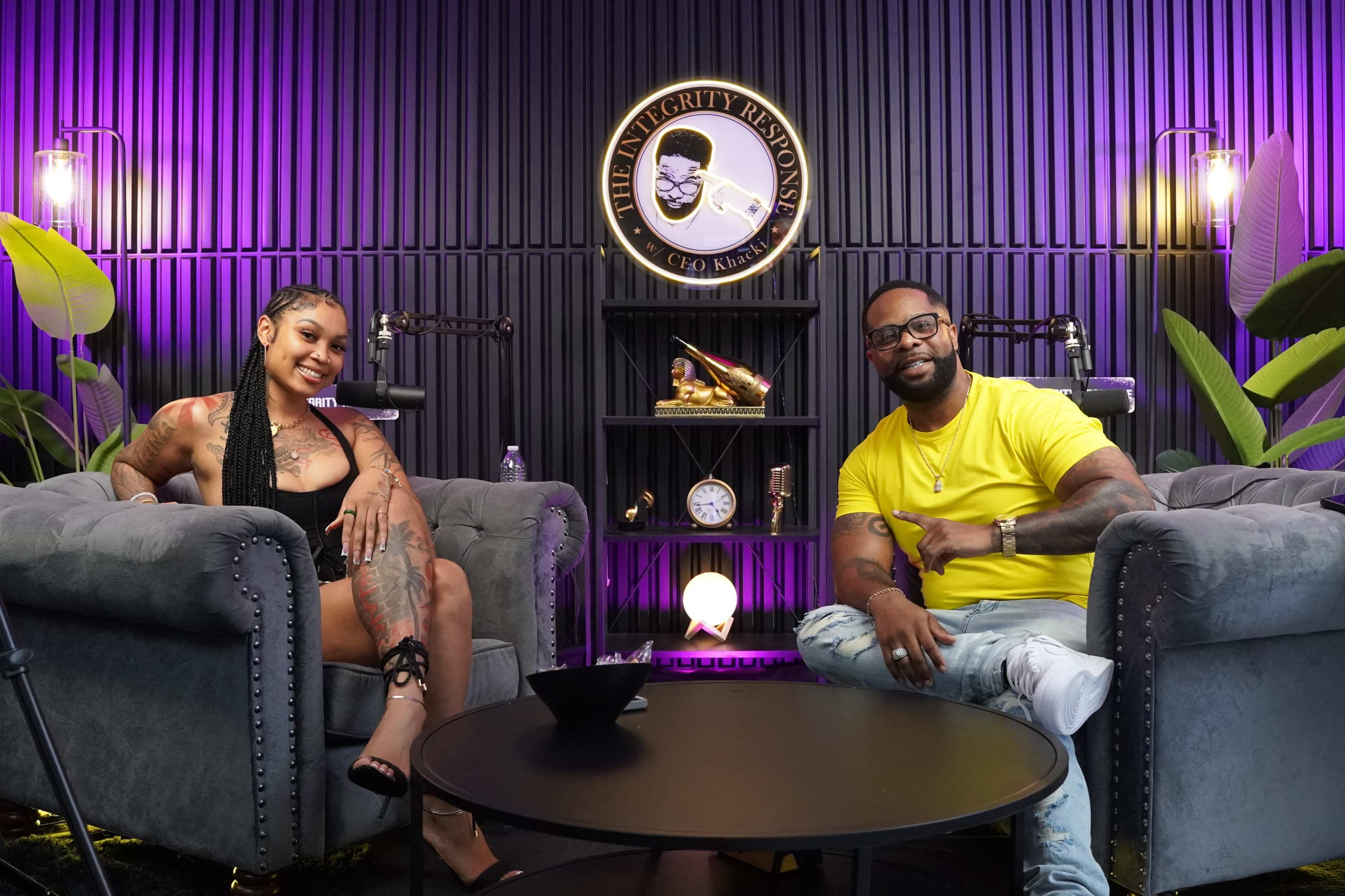 A man and a woman sit in modern armchairs in a vibrant room decorated with plants and a circular sign featuring a face.
