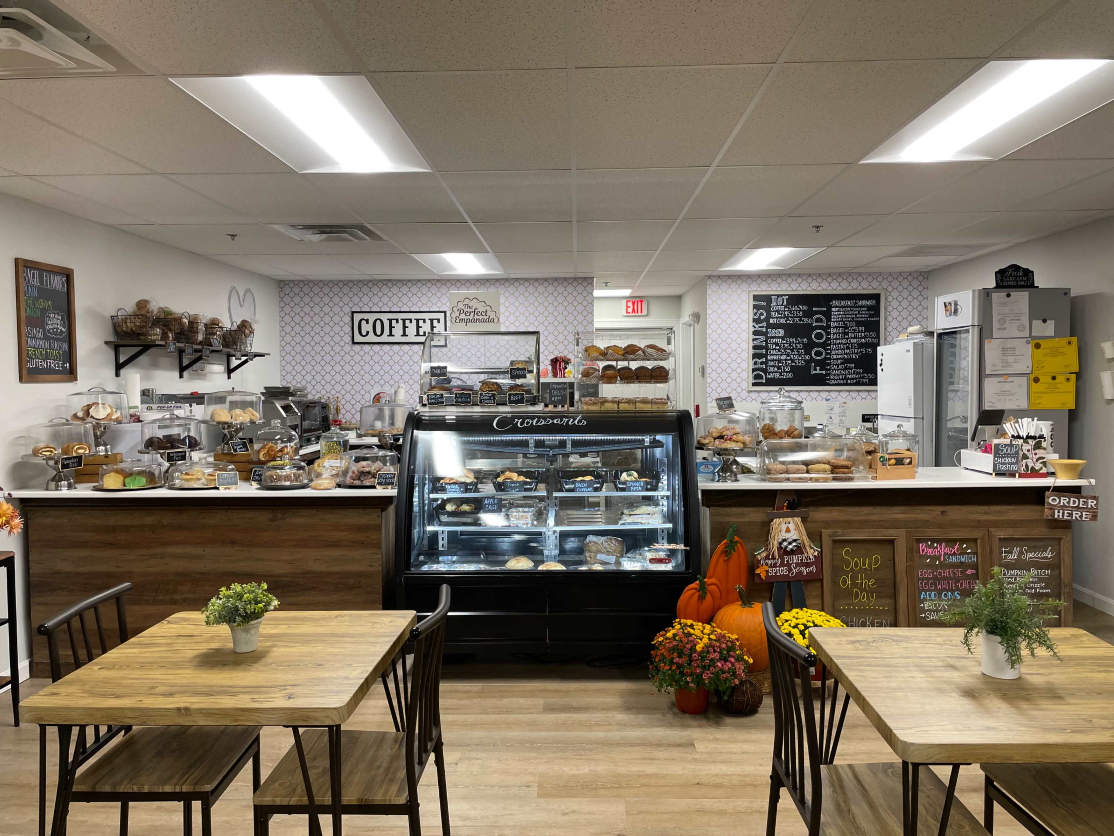 A cozy café features a display case of baked goods, a coffee station, and wooden tables adorned with small plants and flowers.