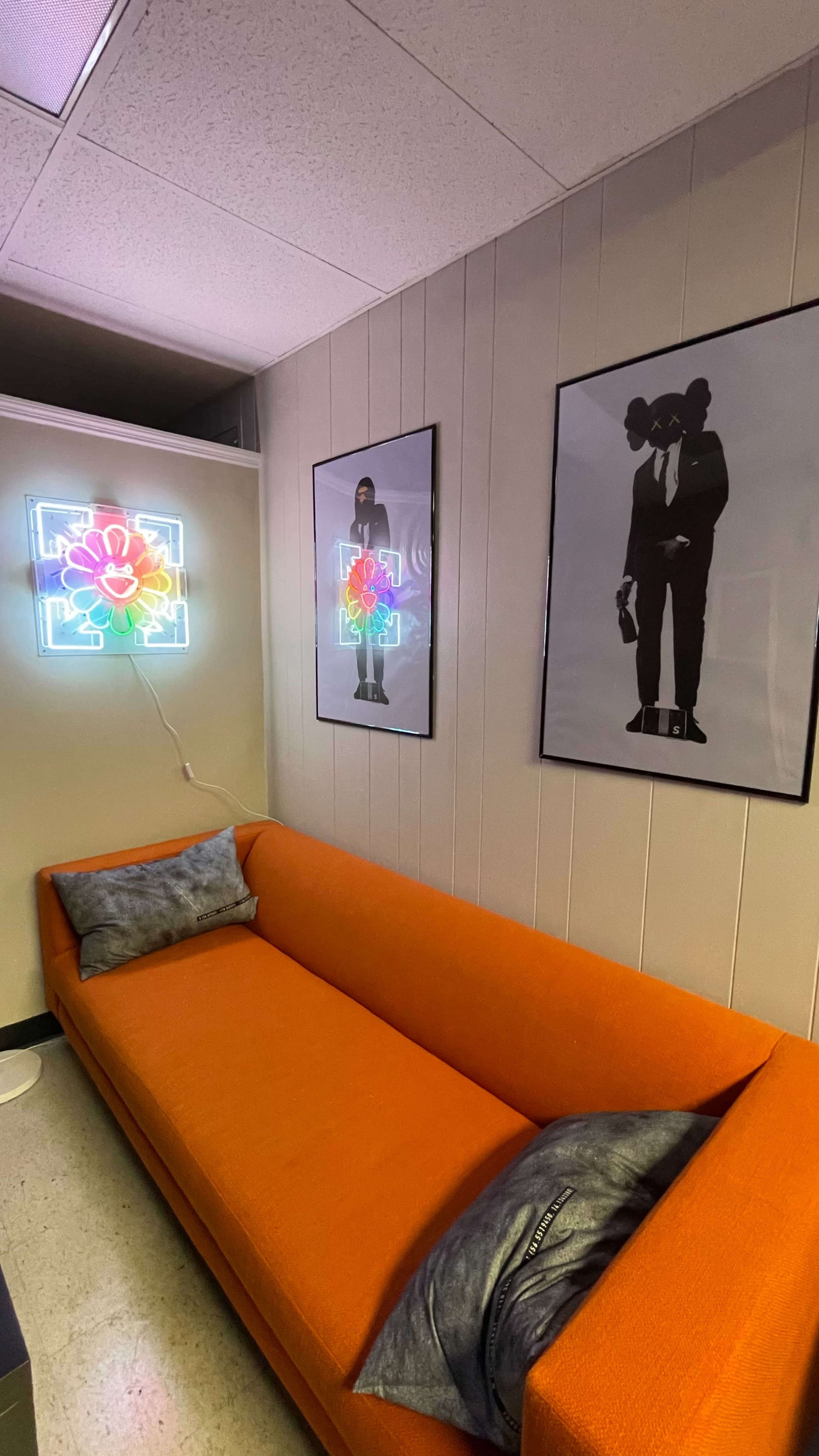 The image shows an orange couch in a corner of a room, flanked by two framed art pieces and a neon flower light on the wall.