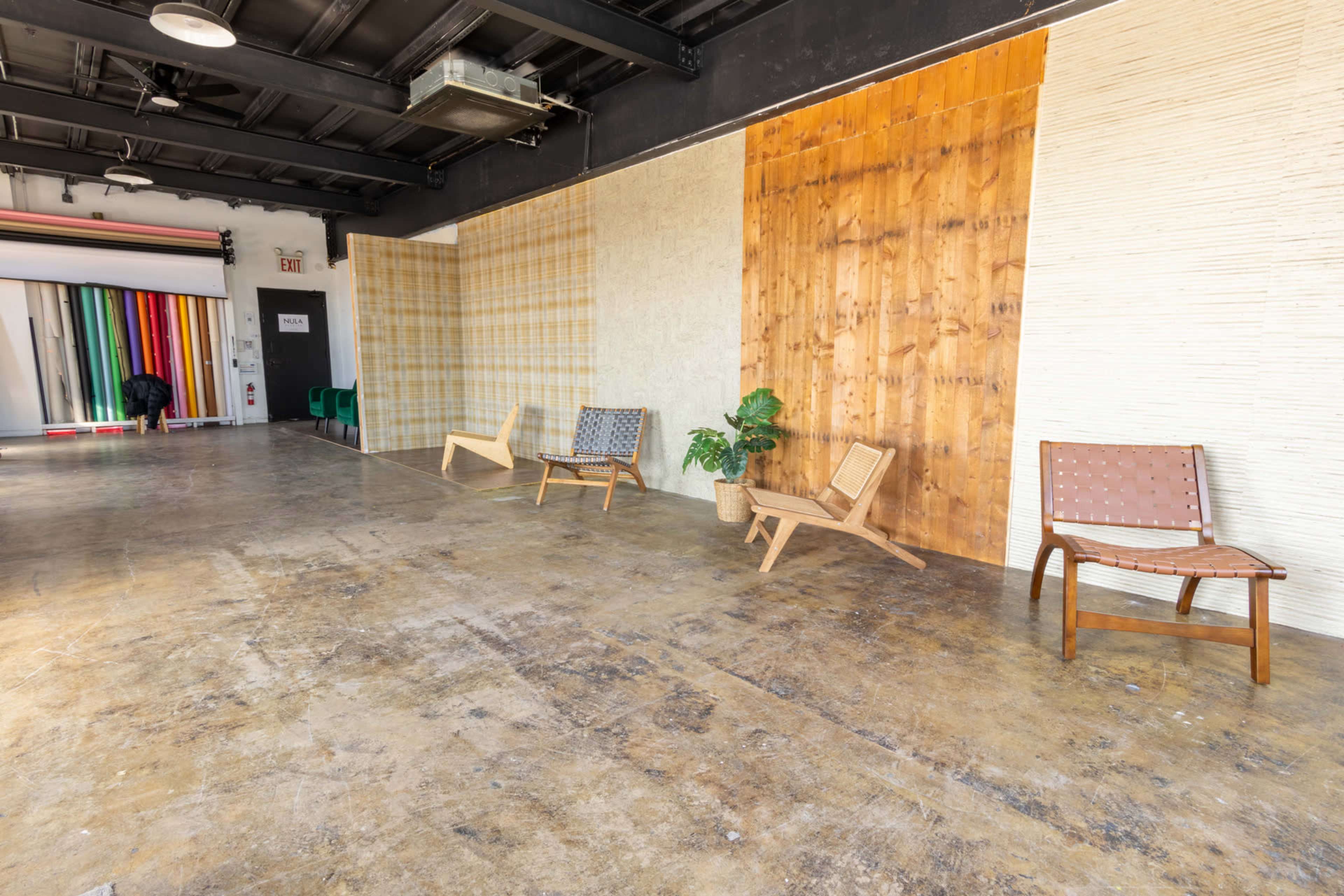The image shows a spacious interior with wooden walls, concrete flooring, and three wooden chairs arranged along one side, alongside a small potted plant.
