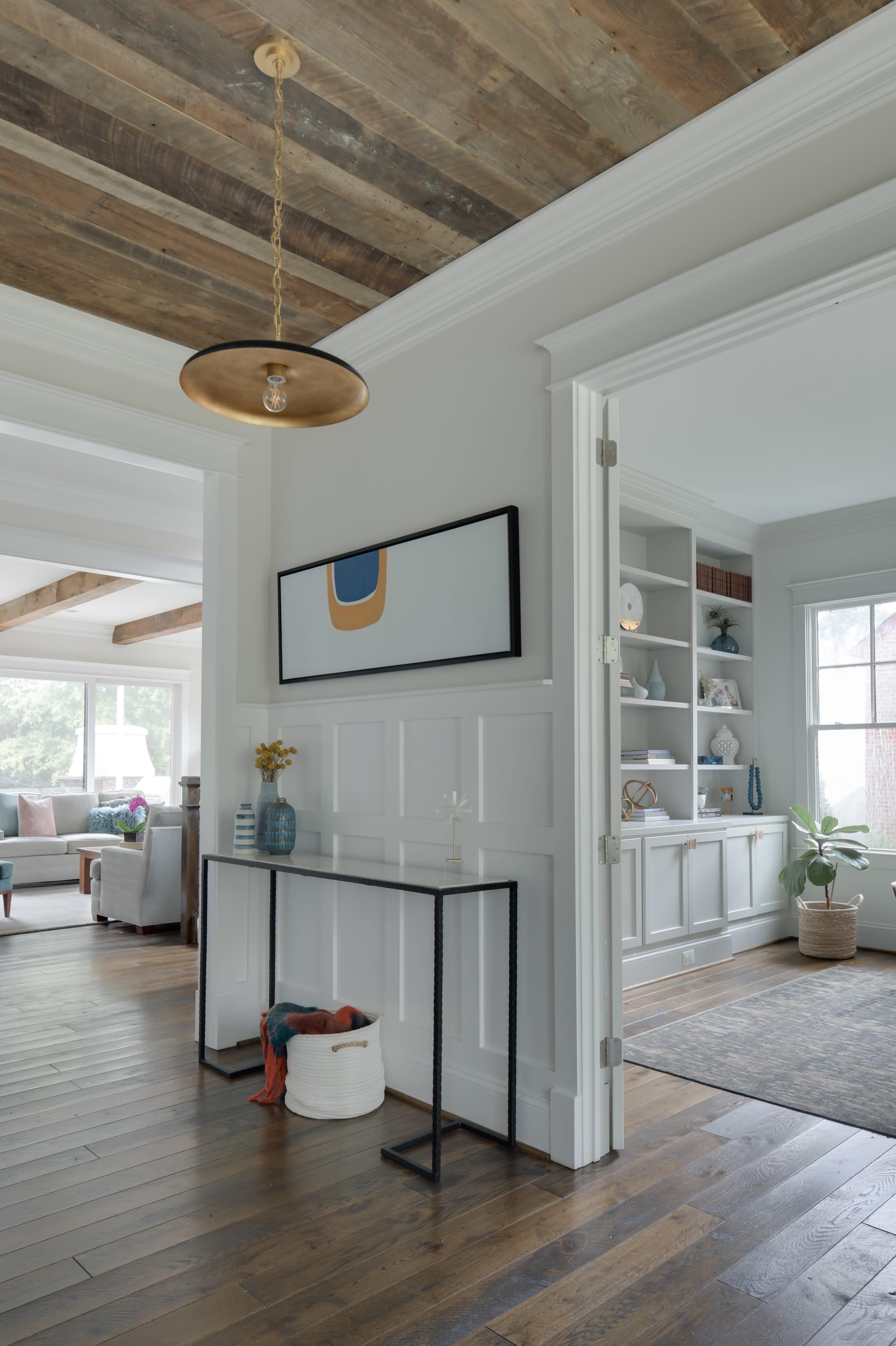 A hallway with a console table, a piece of art on the wall, and an open doorway leading to a living area with large windows.