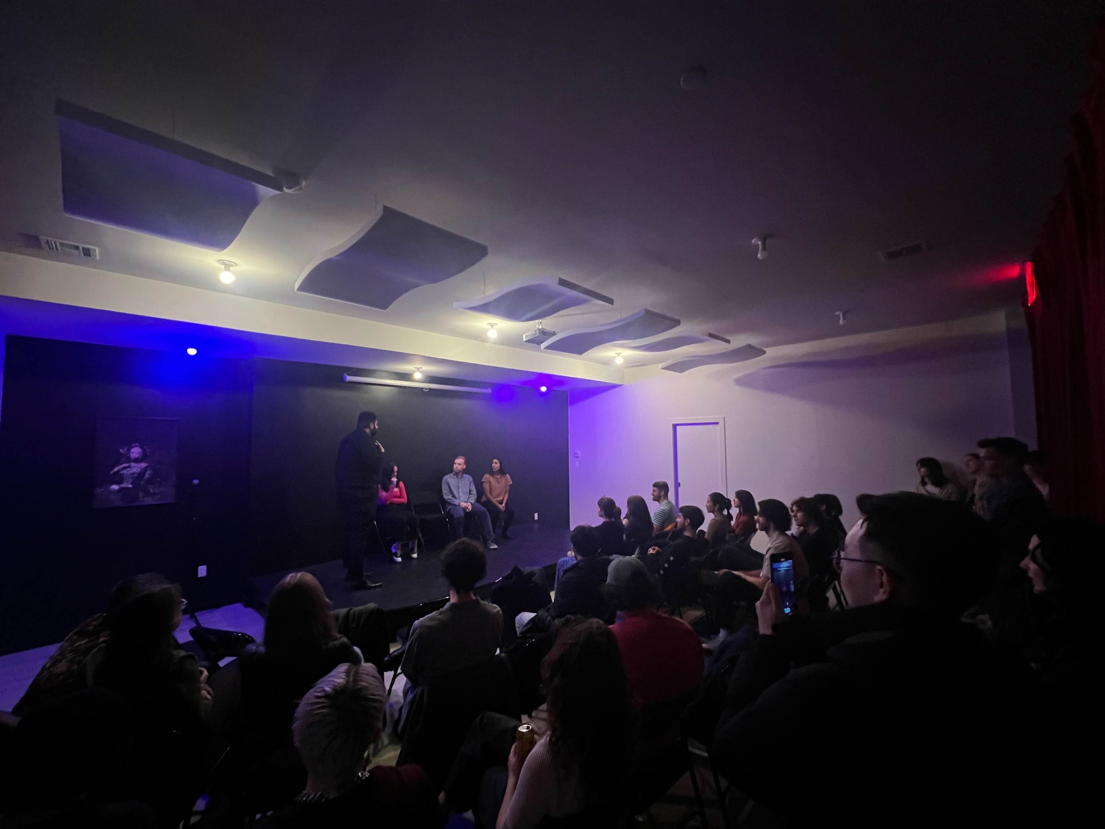 A seated audience watches a panel discussion on a small stage illuminated by dim blue lights.