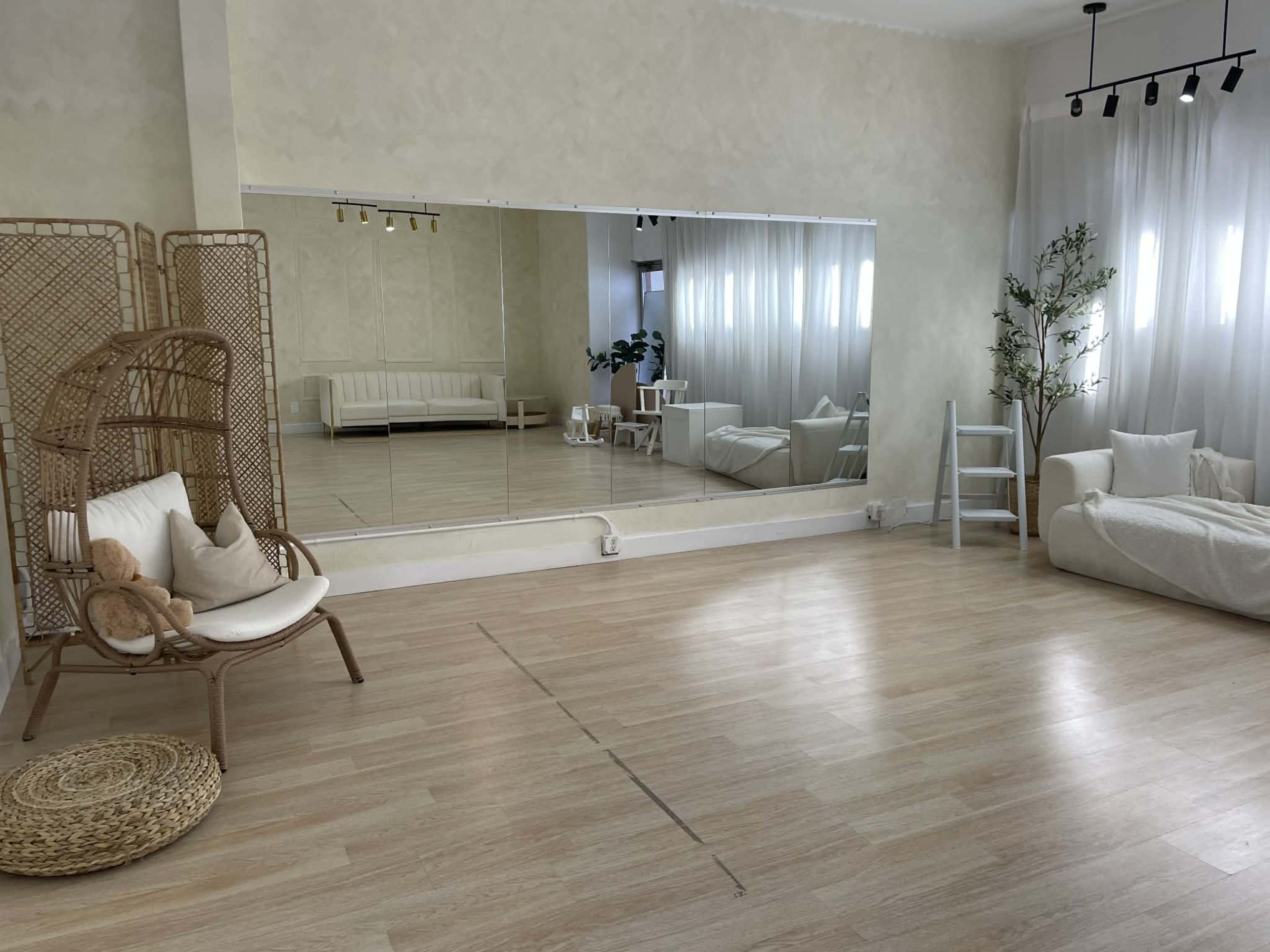 The image shows a bright, spacious room with light-colored walls, wooden flooring, and a large mirror along one side, furnished with a wicker chair, a couch, and potted plants.