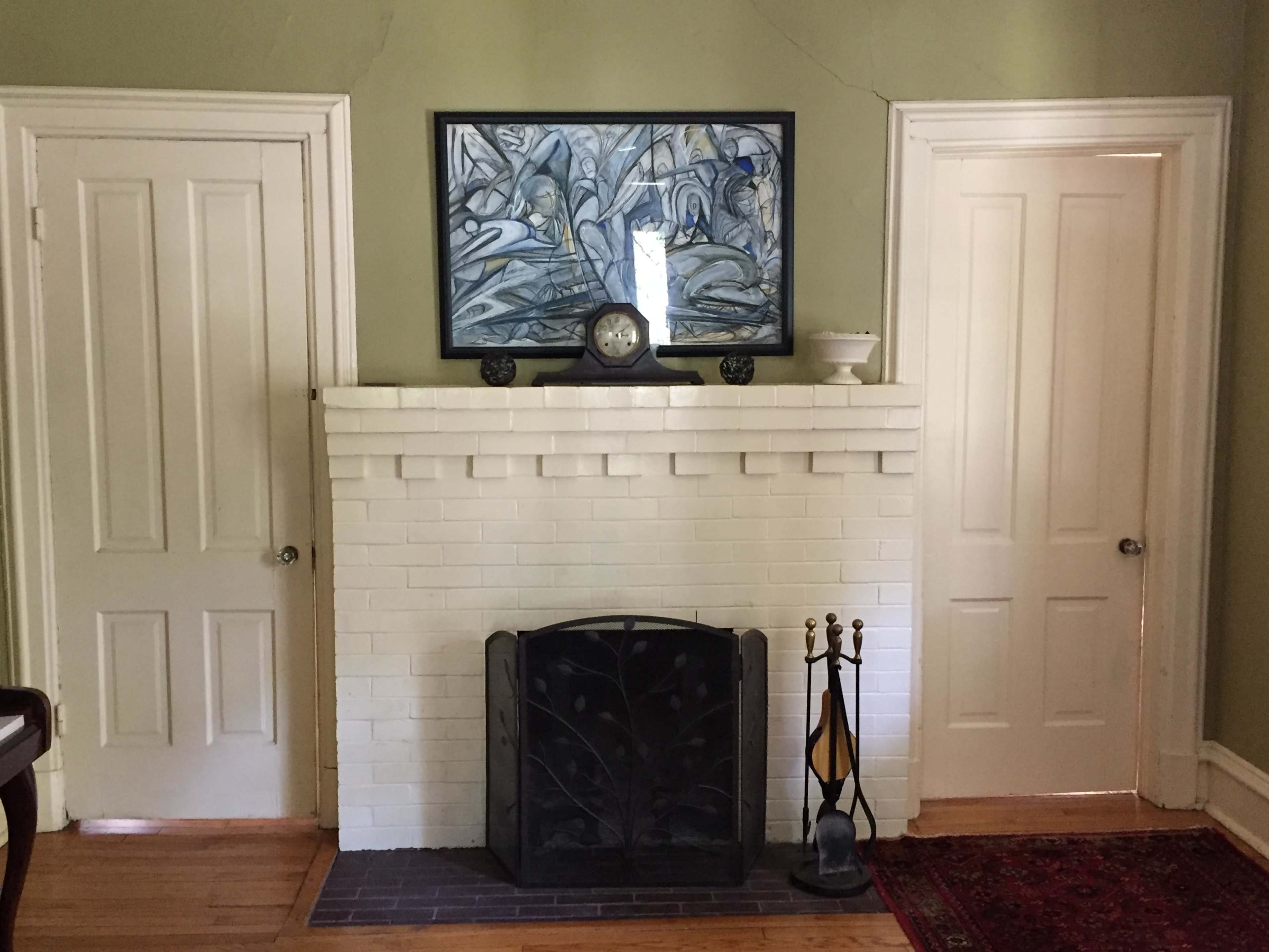 The image shows a white brick fireplace flanked by two closed white doors, with a clock and artwork above the mantel.