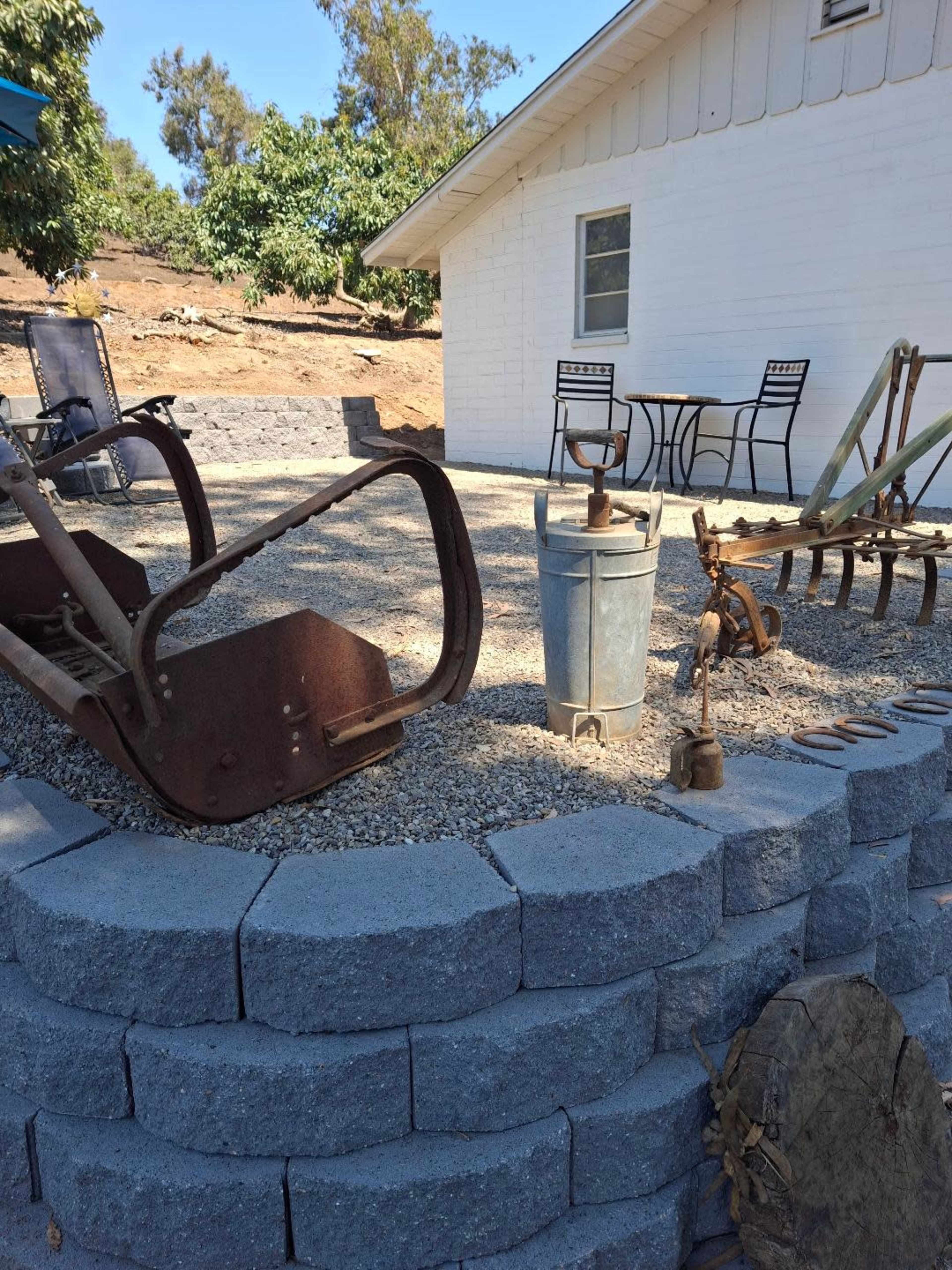 The scene shows a gravel area with a brick retaining wall, featuring various rustic farming tools and equipment placed nearby a white house and a small outdoor table with chairs.
