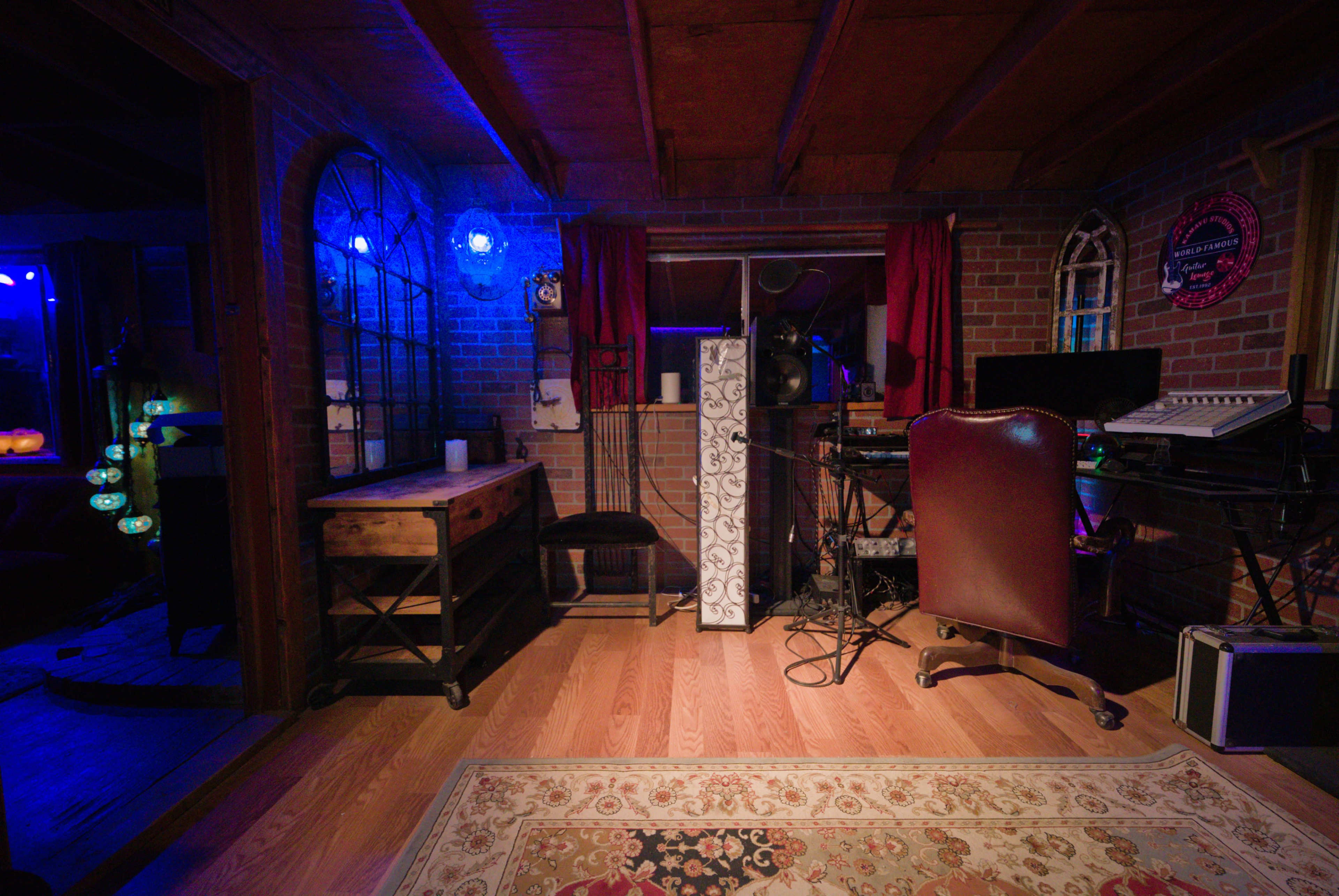 The image shows a cozy workspace with a desk, a chair, and various electronic equipment set against a backdrop of exposed brick walls and wooden beams.