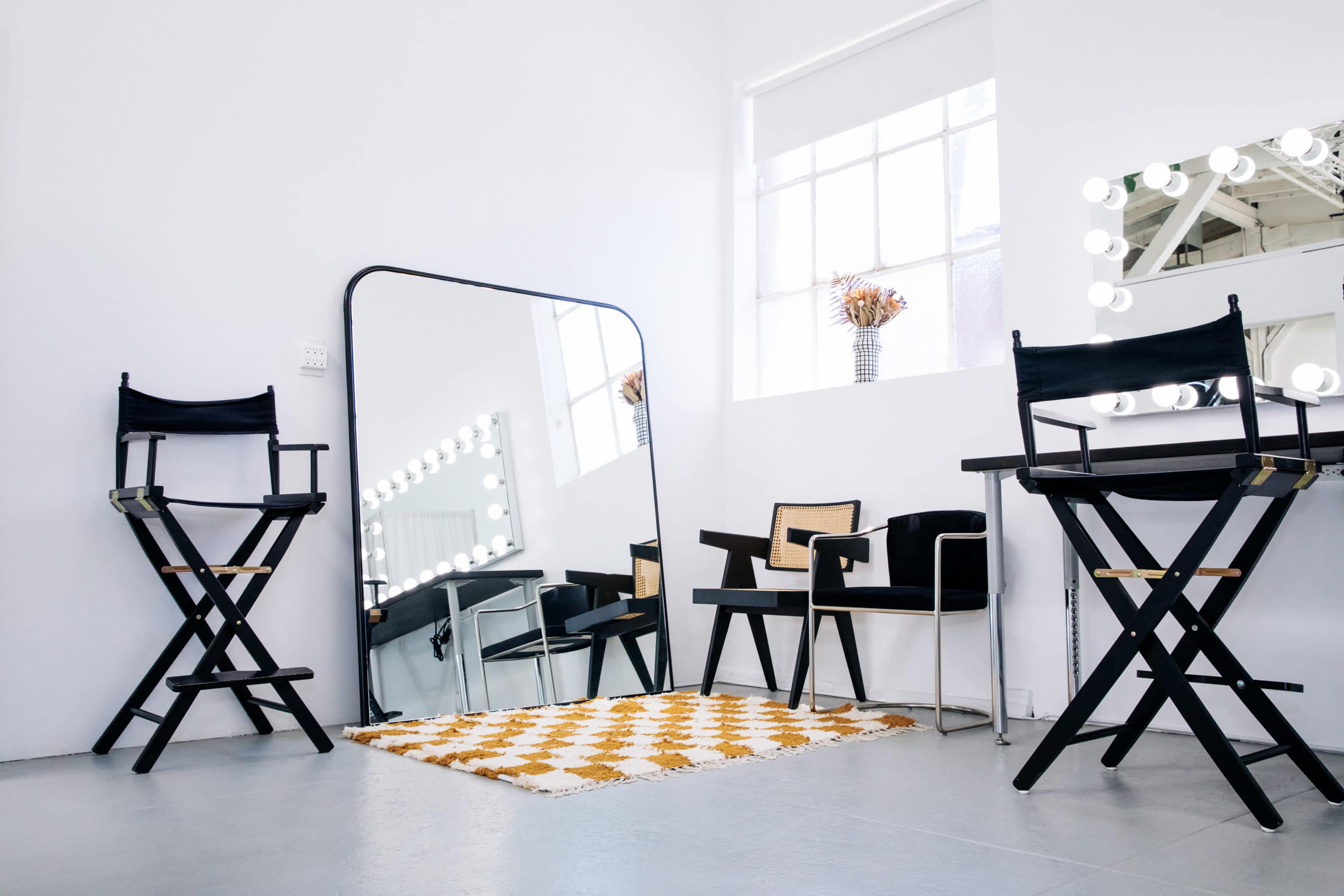 A bright, modern salon interior featuring two black director's chairs, a large mirror, and a table with a lighted mirror backdrop.