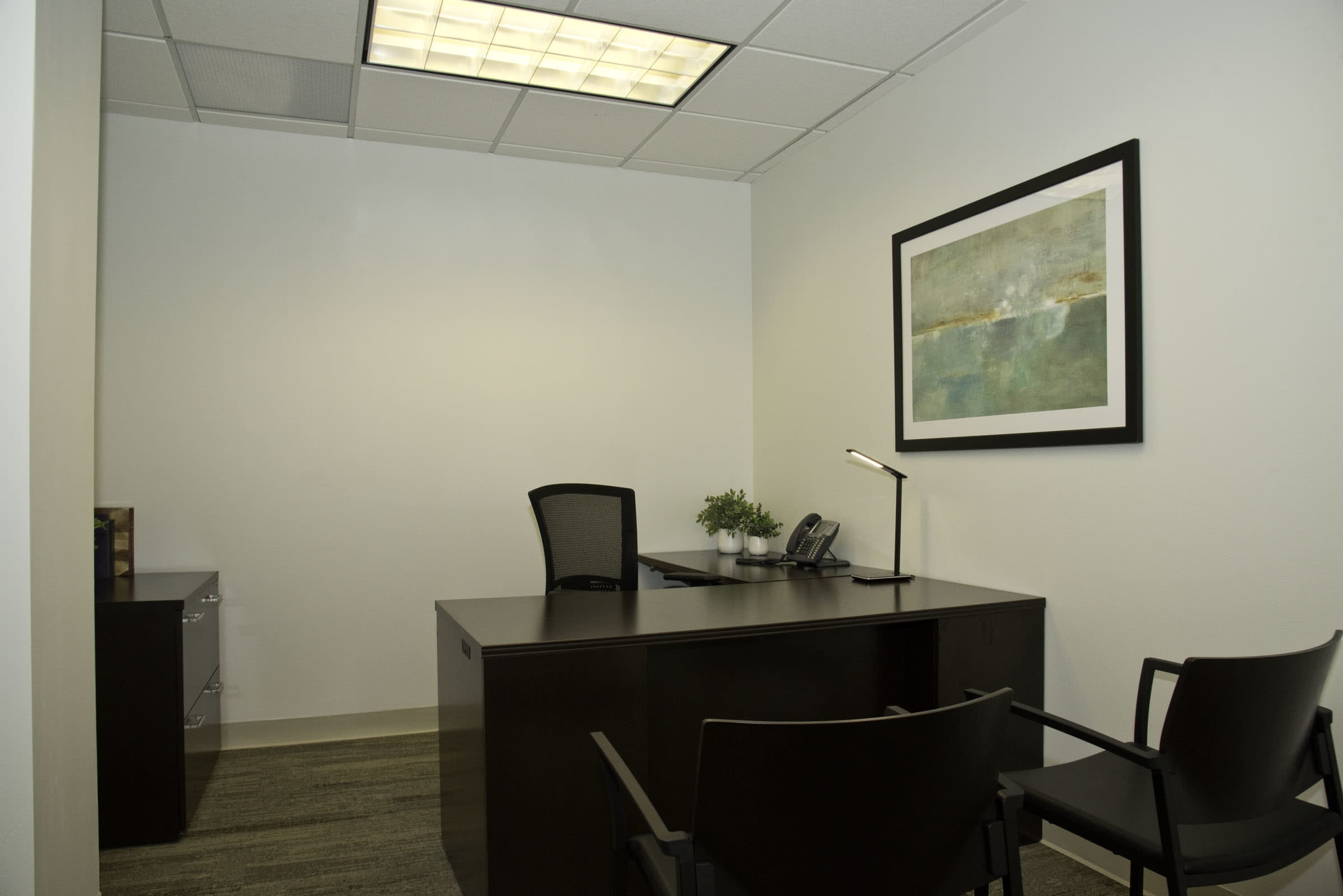 The image shows a minimalist office space with a desk, two chairs, a phone, and a wall art piece.