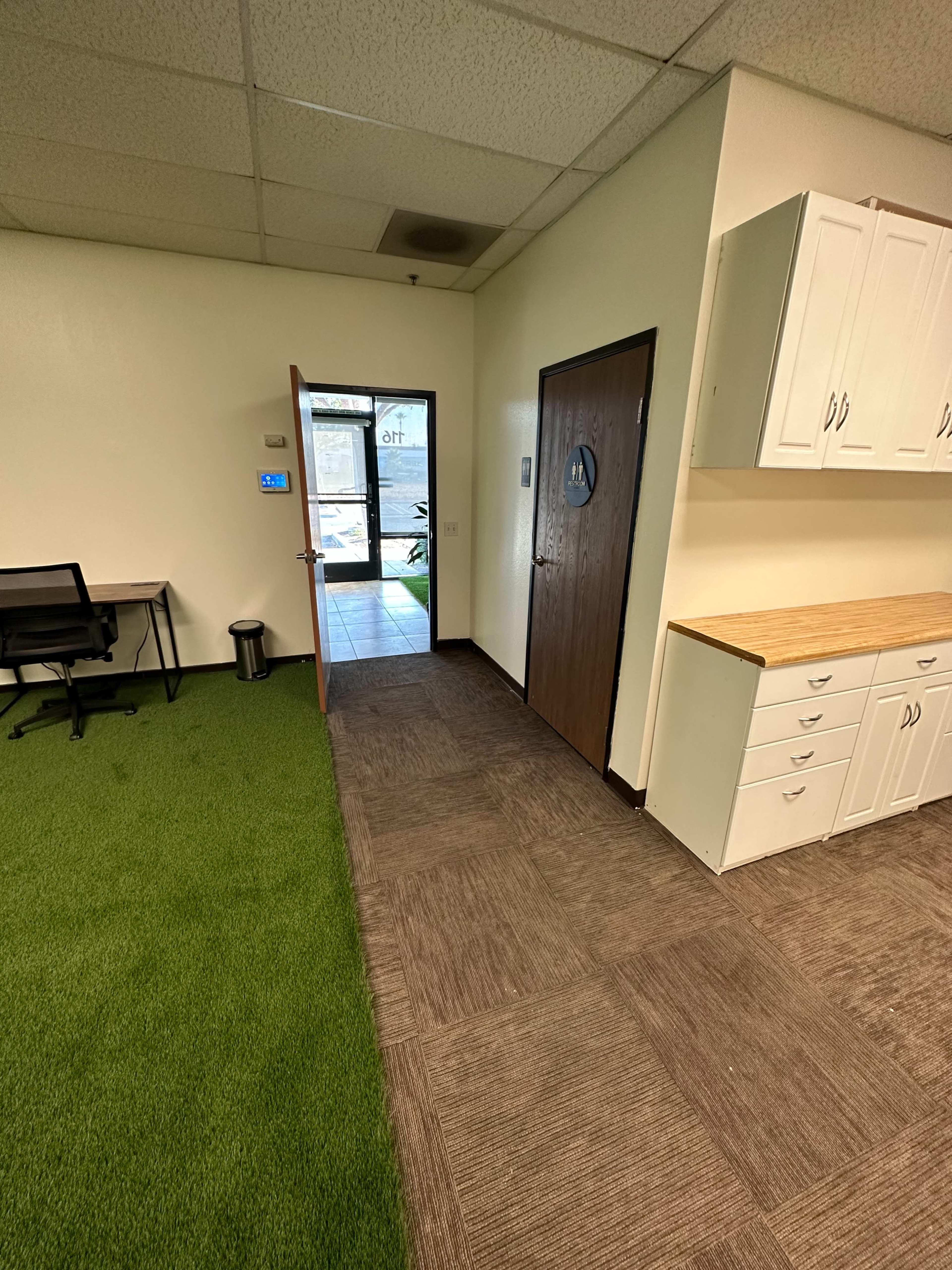 An office space featuring a doorway leading outside, a chair at a desk, and tiled flooring adjacent to green carpet.