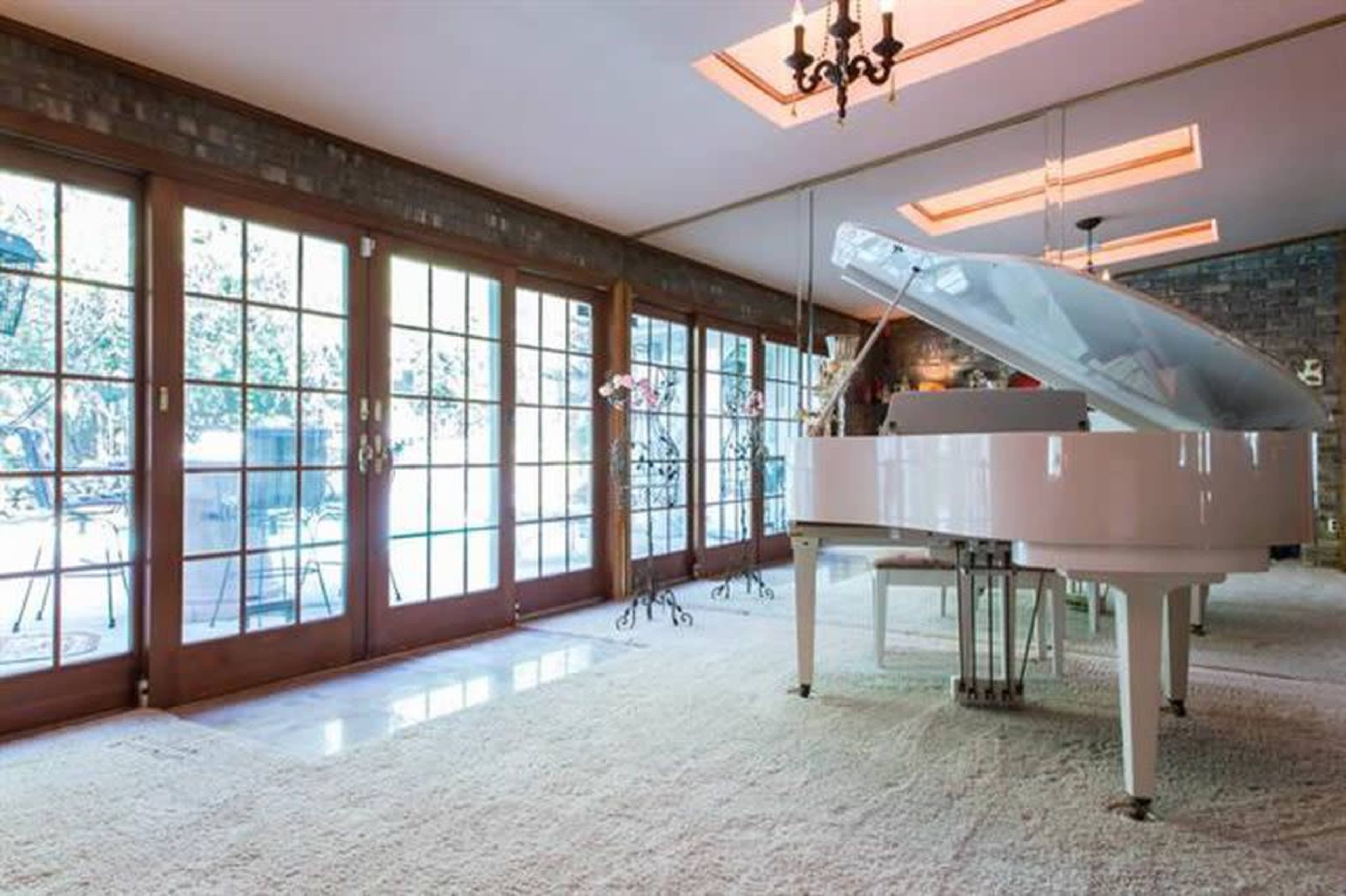 A white grand piano is positioned on a carpeted floor in front of large glass doors that open to a patio area.