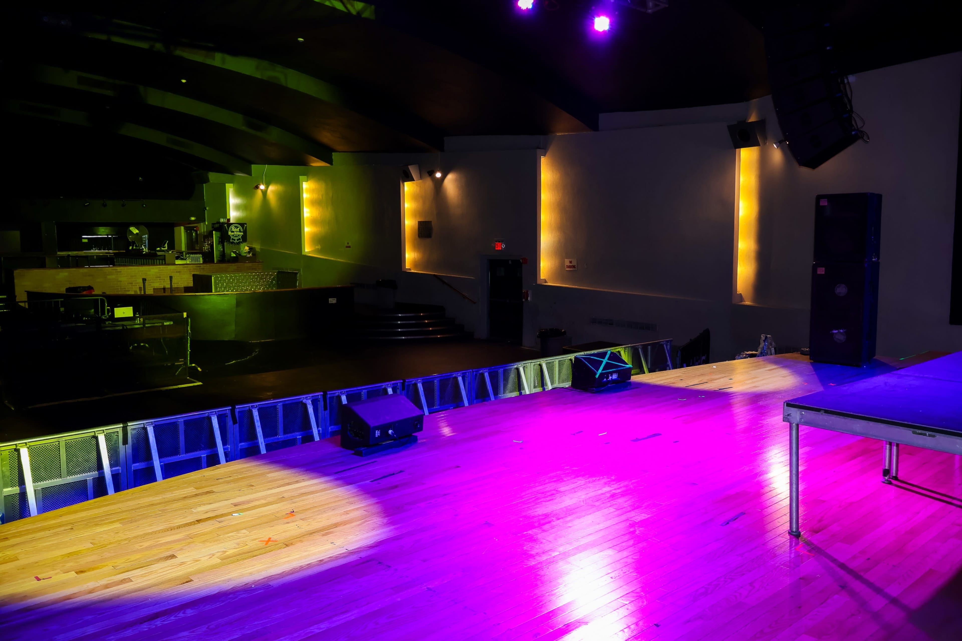 A stage with purple lighting and sound equipment is set up in a dimly lit venue, showcasing polished wooden flooring and a raised seating area in the background.