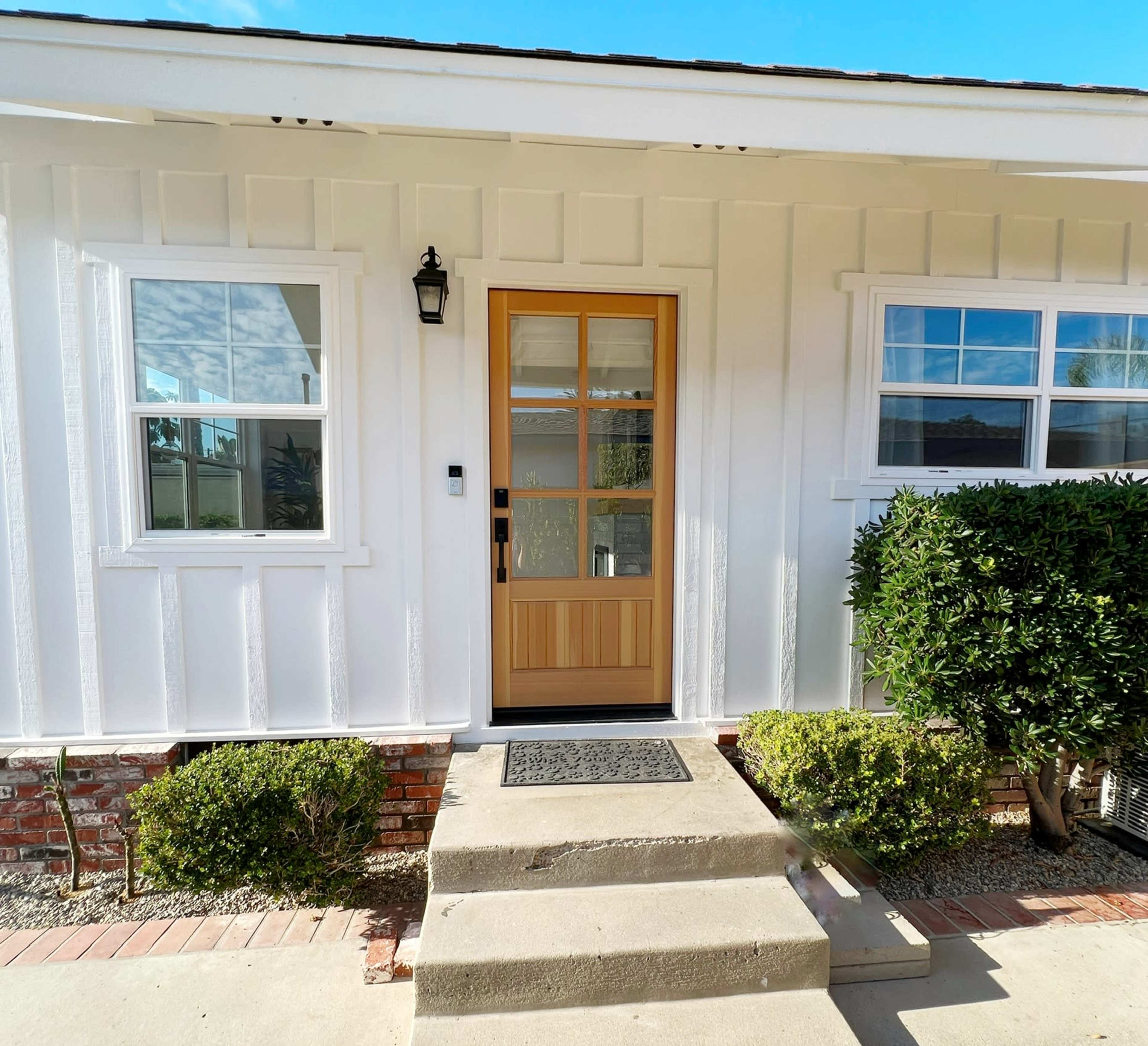 A front door made of wood, framed by two windows, situated on a white house with a concrete step and landscaped bushes.