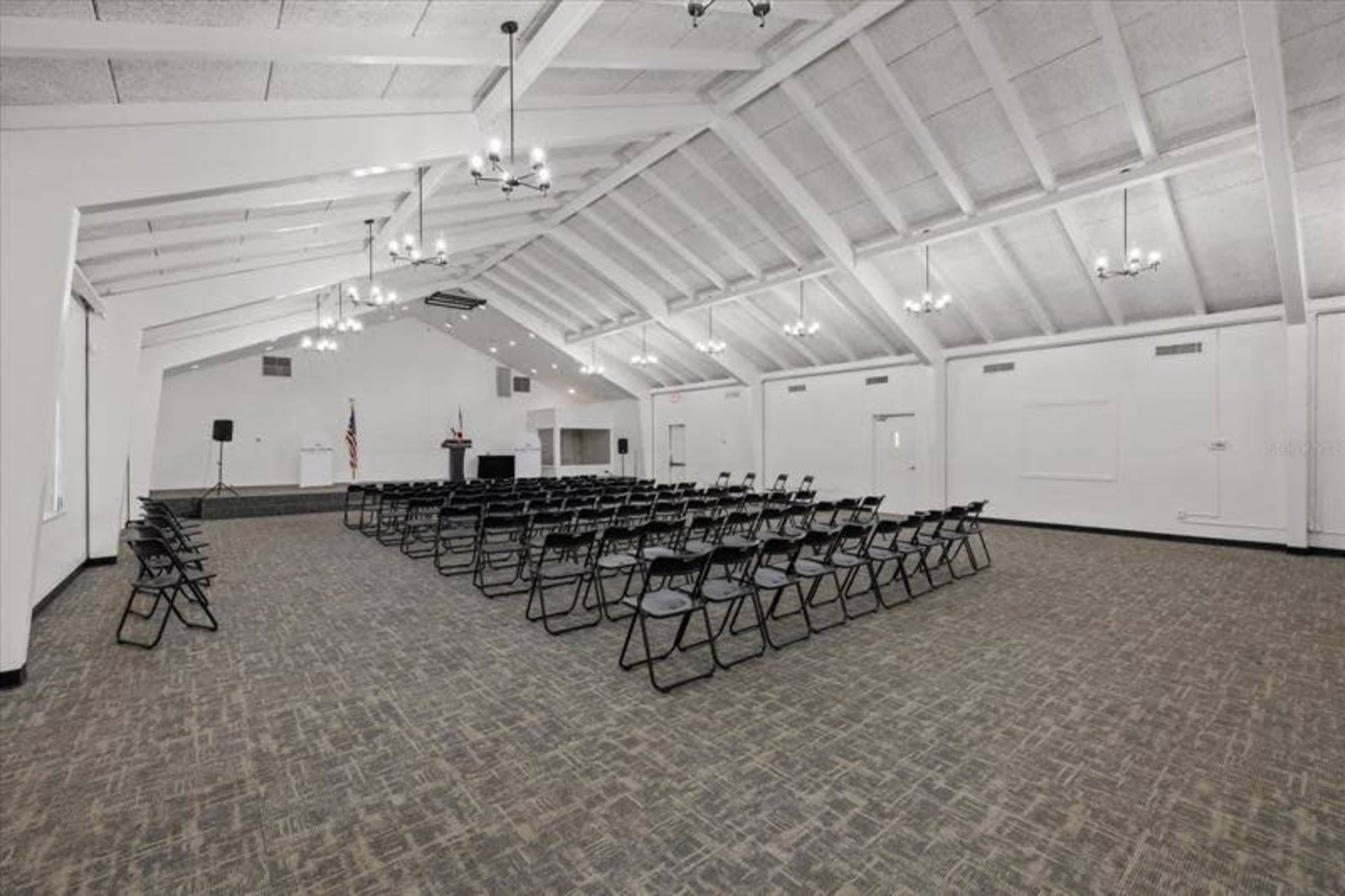 The image shows a spacious, well-lit hall arranged with rows of black folding chairs facing a podium and an American flag.