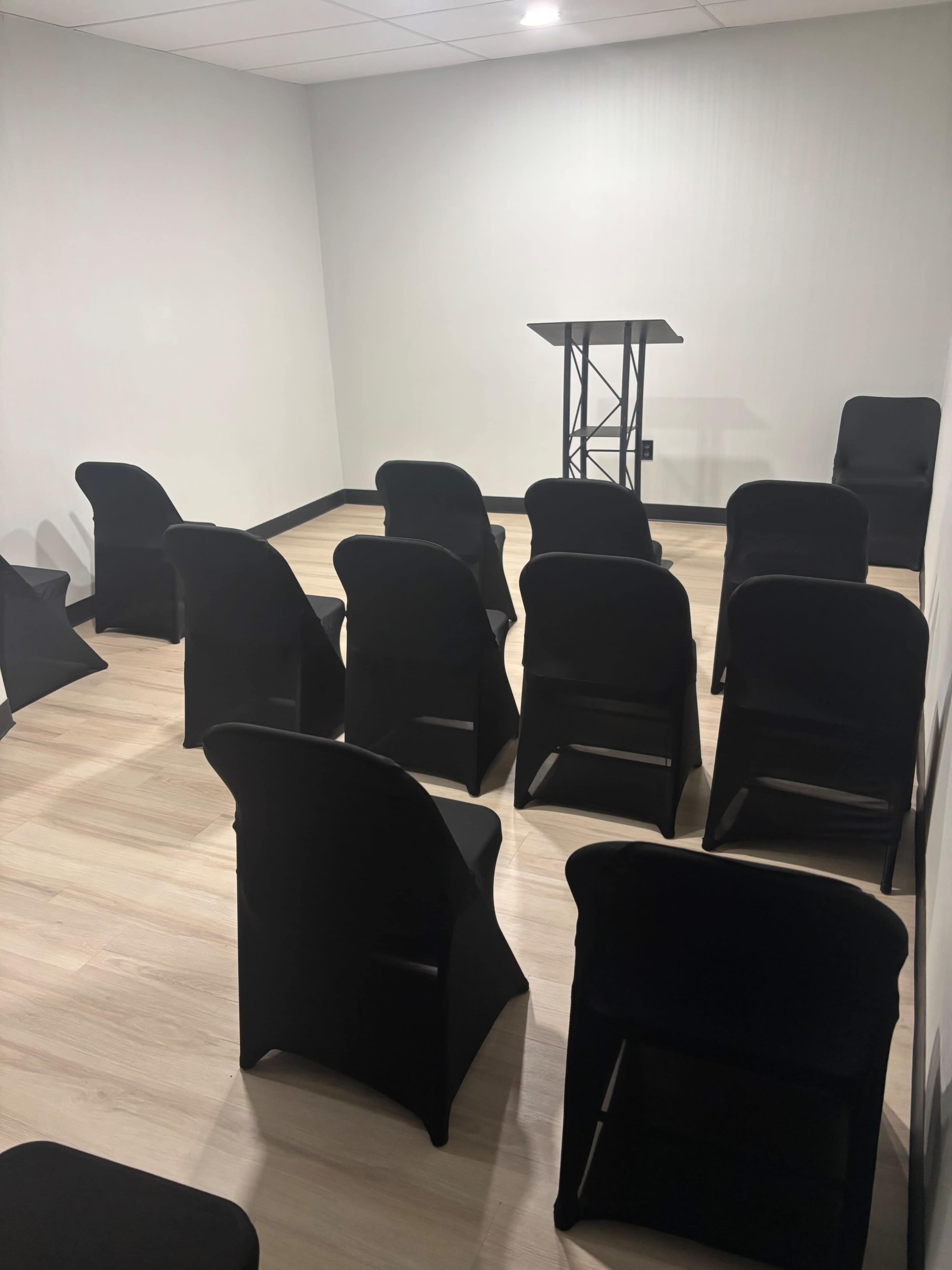 The image shows a small, empty room with black chairs arranged in rows facing a raised podium.