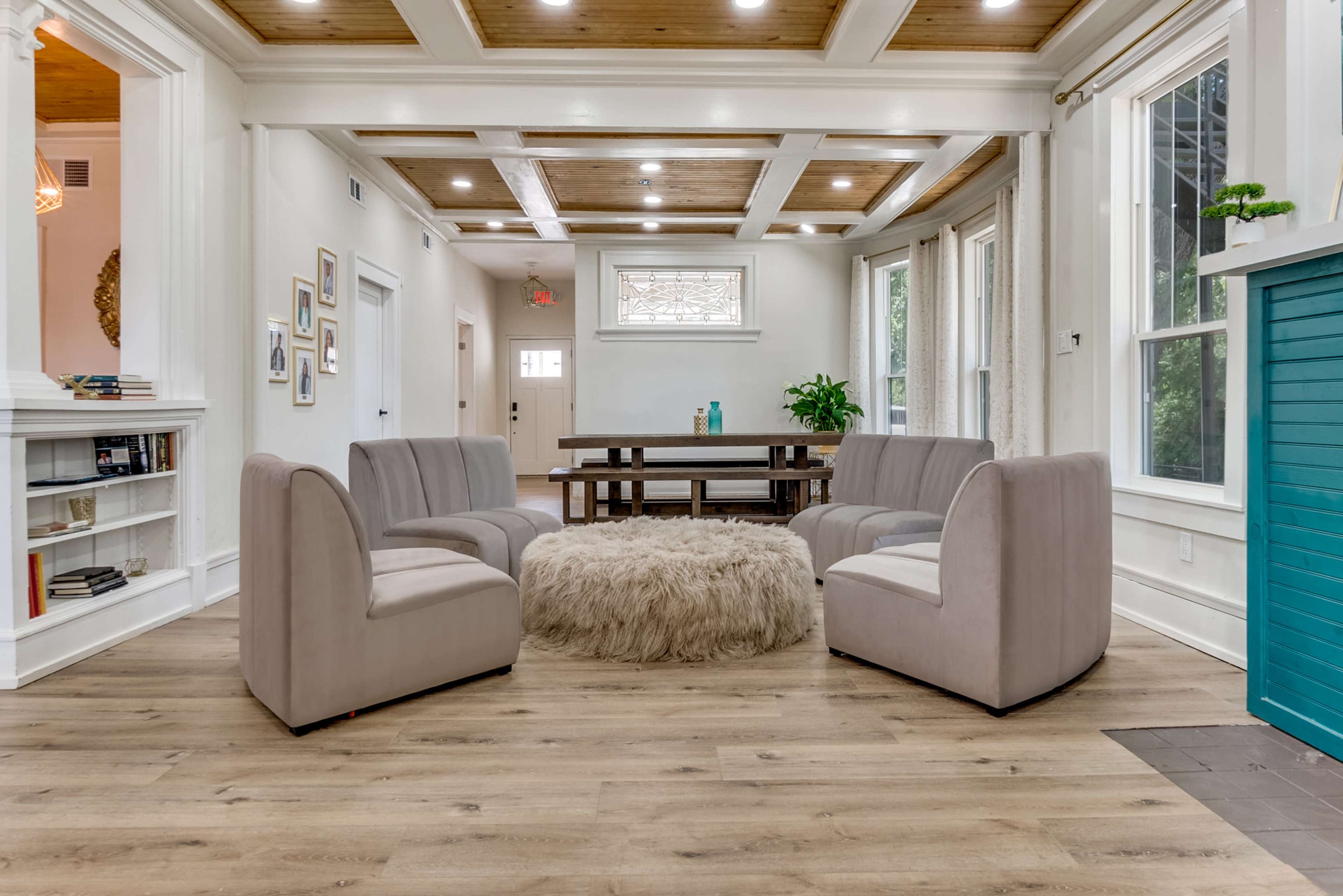 The image shows a spacious, well-lit room with a circular arrangement of six beige armchairs around a round, fluffy area rug, accompanied by a large wooden dining table and decorative plants.