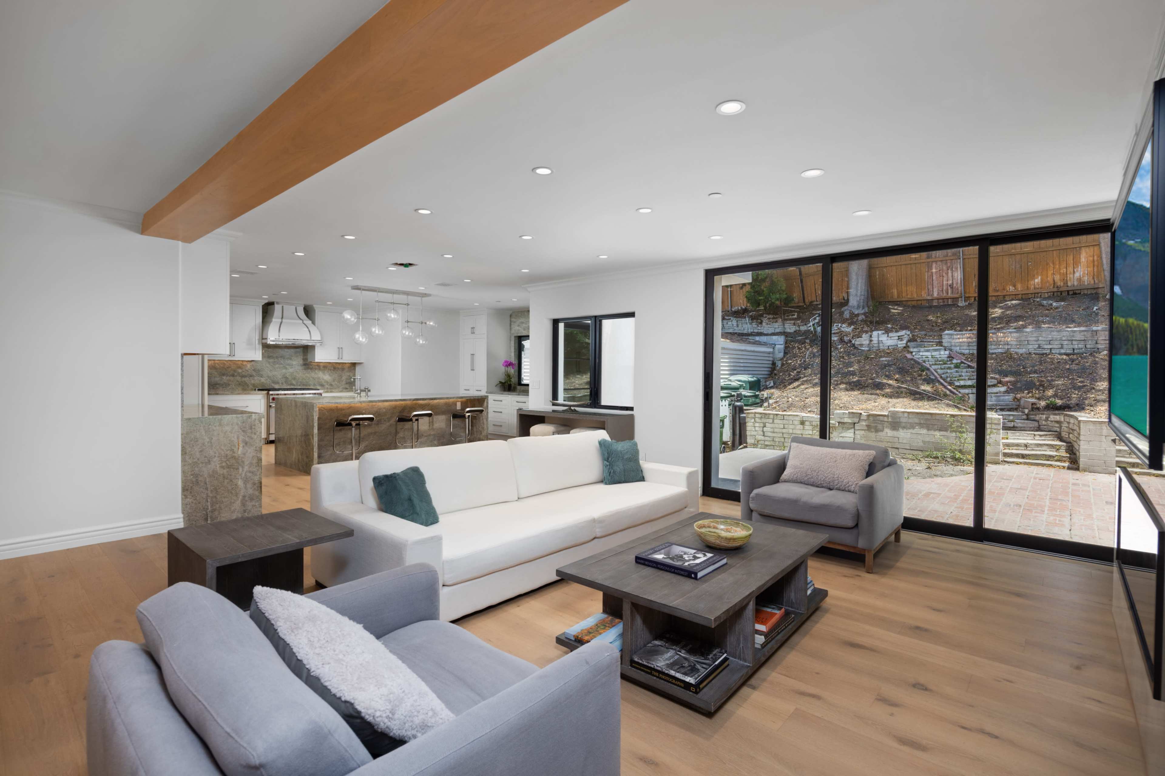 A modern living room with a white sofa and gray armchairs, featuring large windows that overlook a patio and garden area.