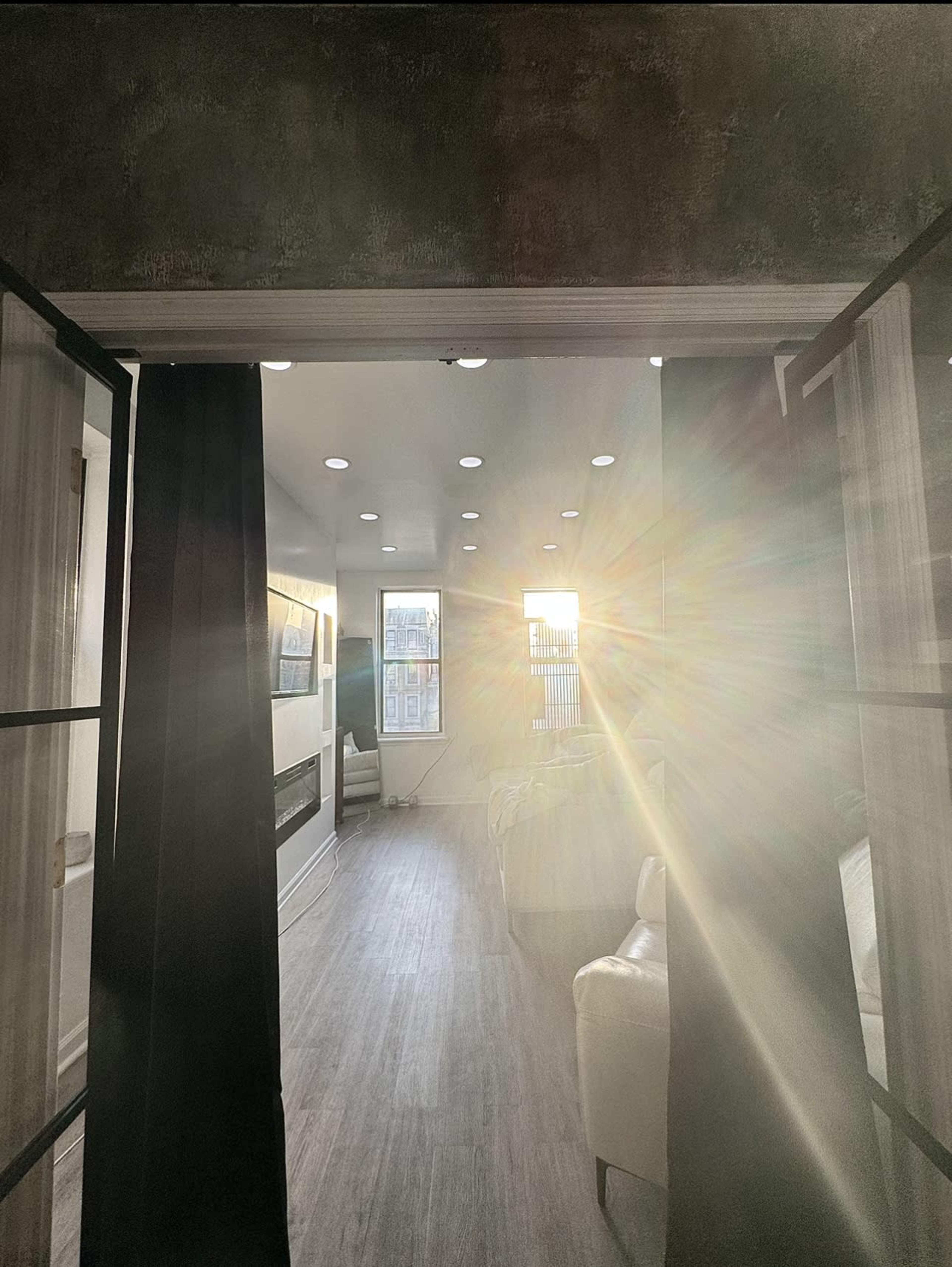 A hallway with black curtains leads to a brightly lit living space, with sunlight streaming through a large window.