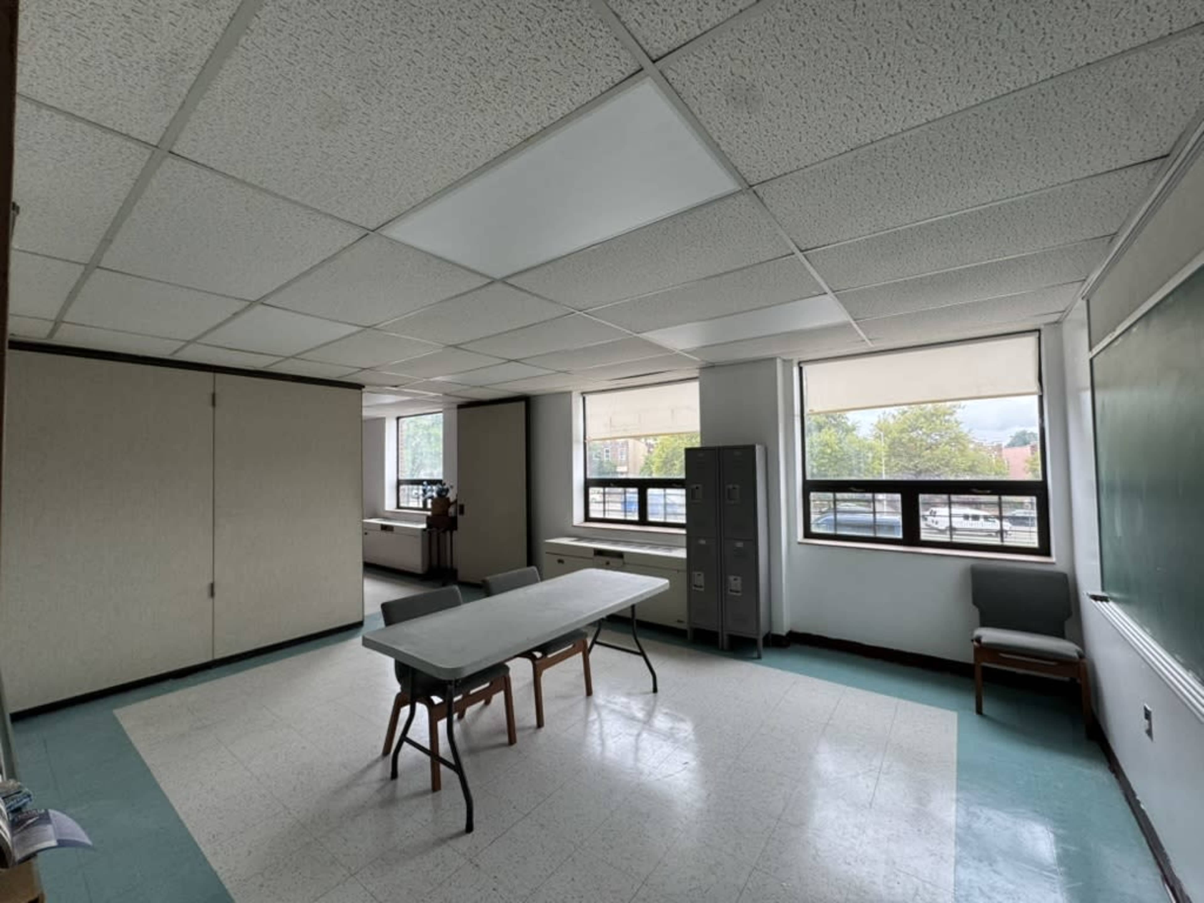 The image shows a classroom with a table and chairs in the center, large windows on two sides, and a chalkboard on one wall.