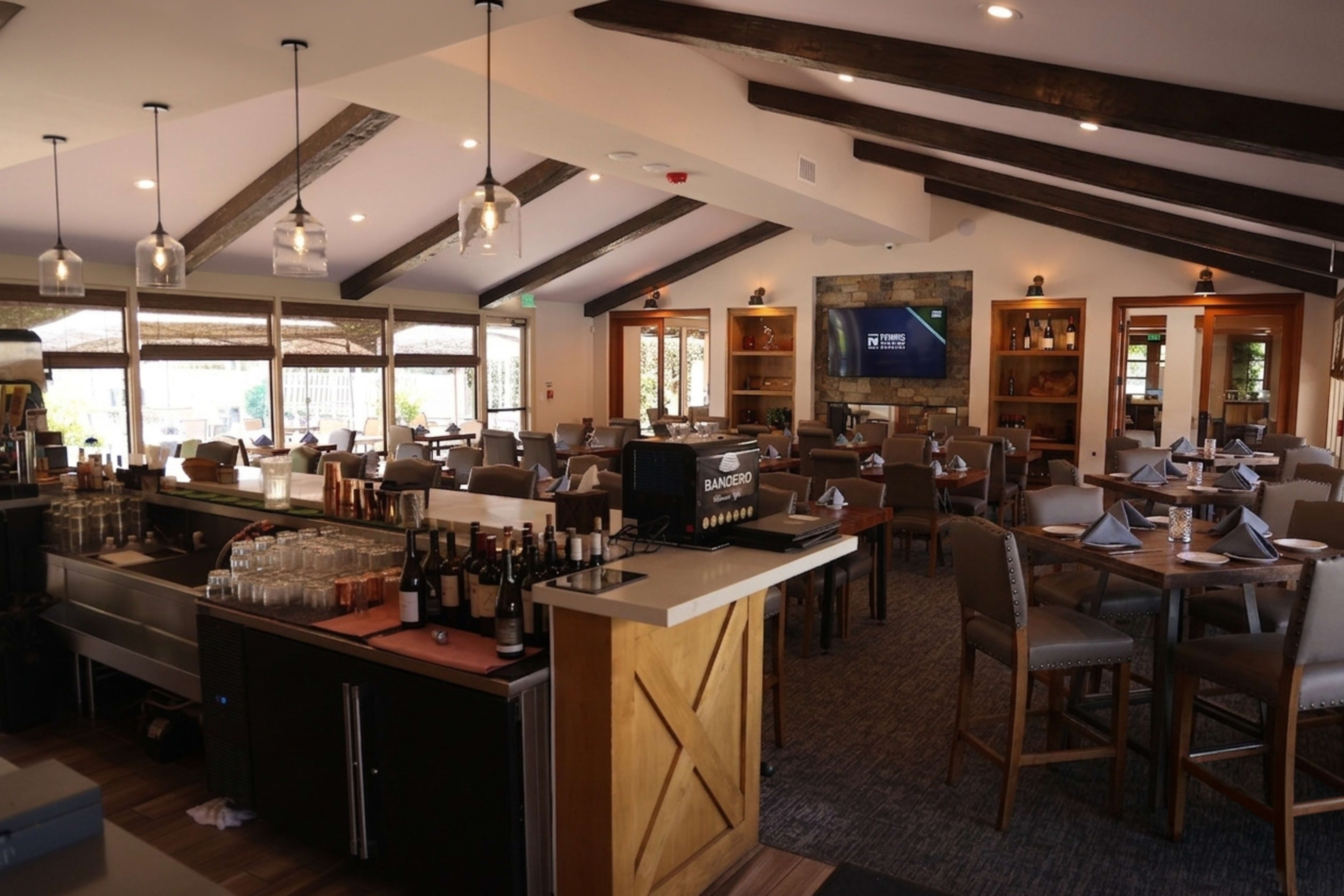 The image shows an interior of a restaurant featuring wooden beams, a bar area with wine bottles, and neatly arranged tables and chairs.