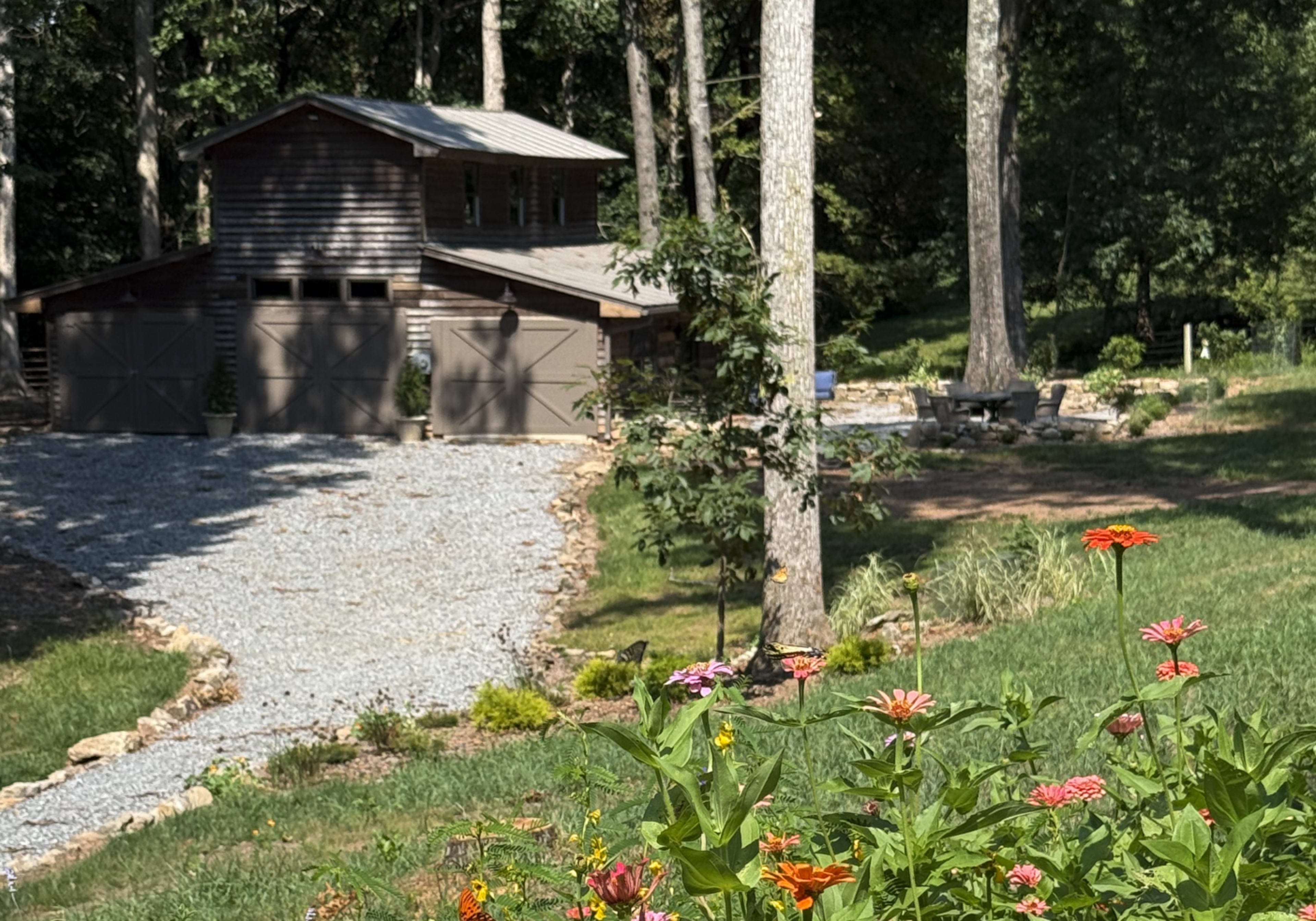 A gravel driveway leads to a wooden garage surrounded by trees, with a garden of colorful flowers in the foreground.