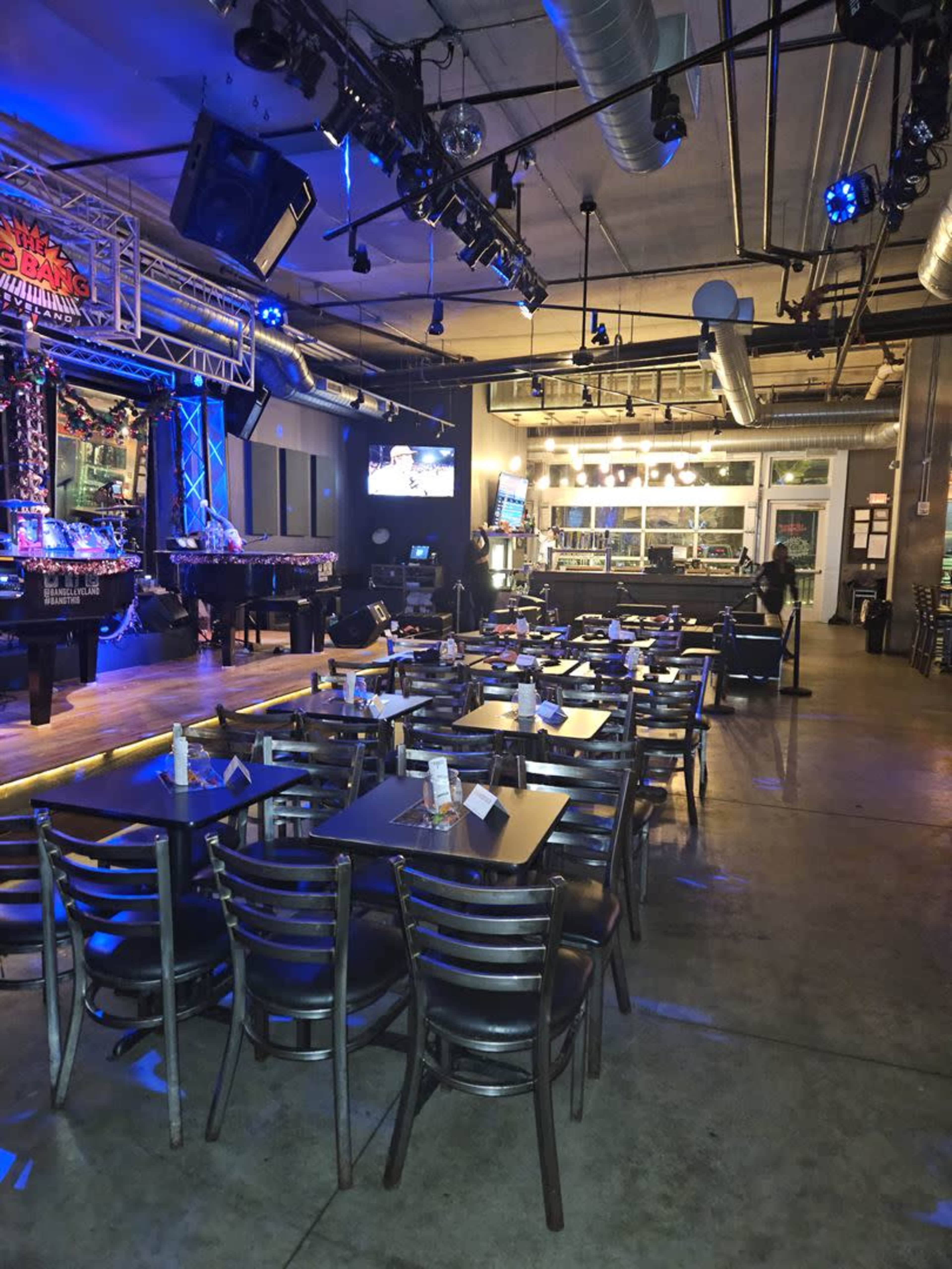 A spacious entertainment venue with rows of black tables and chairs arranged in front of a stage, illuminated by blue lighting.