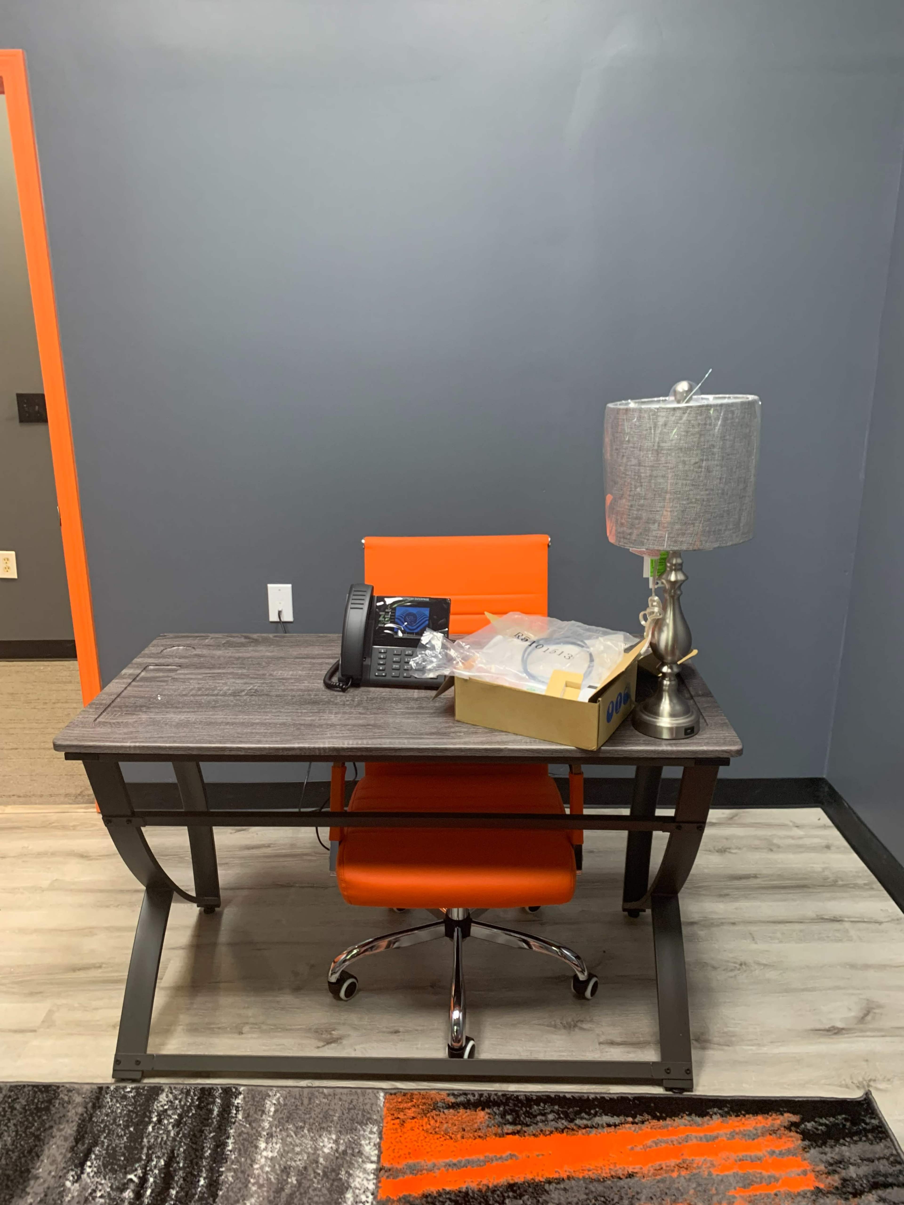A modern office desk with an orange chair, a telephone, and a box on top, set against a gray wall.