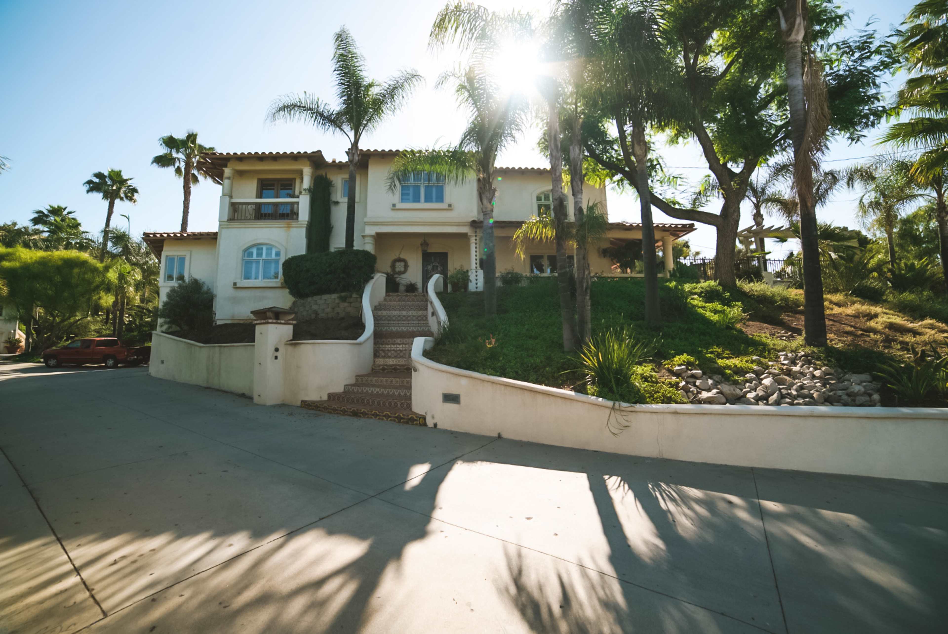 A large two-story house with palm trees and a landscaped yard is positioned on a sloped driveway under bright sunlight.