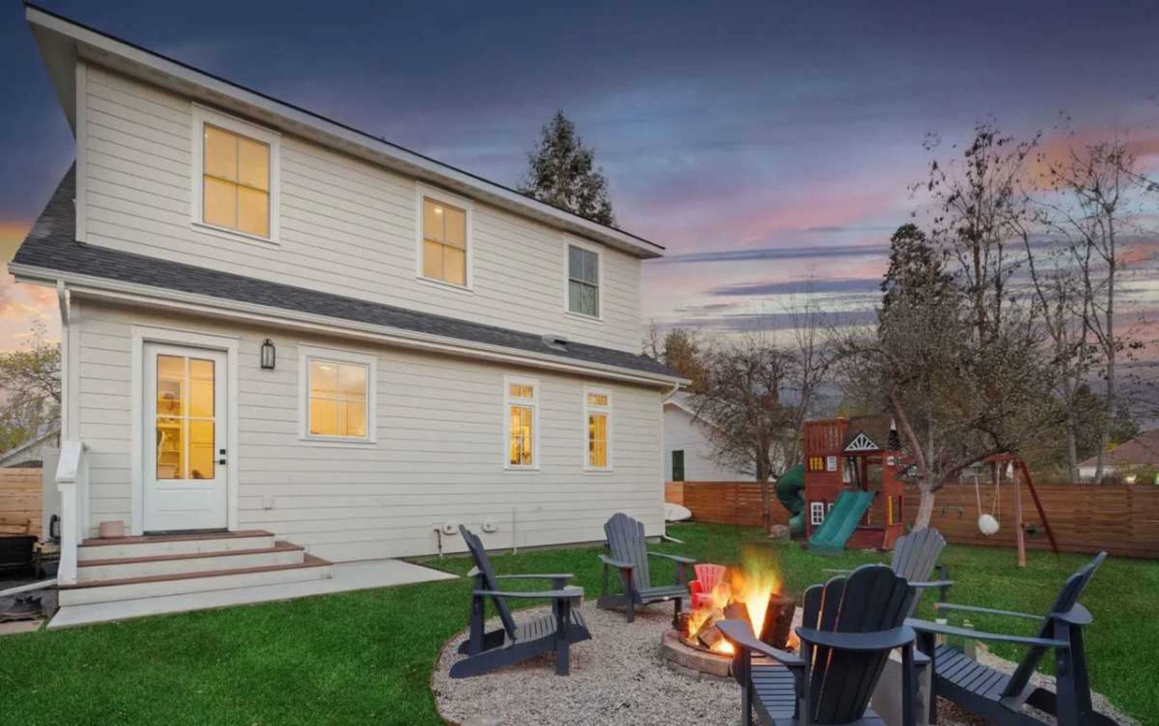 A two-story house with white siding features a fire pit surrounded by chairs in a backyard, with playground equipment visible in the background during sunset.