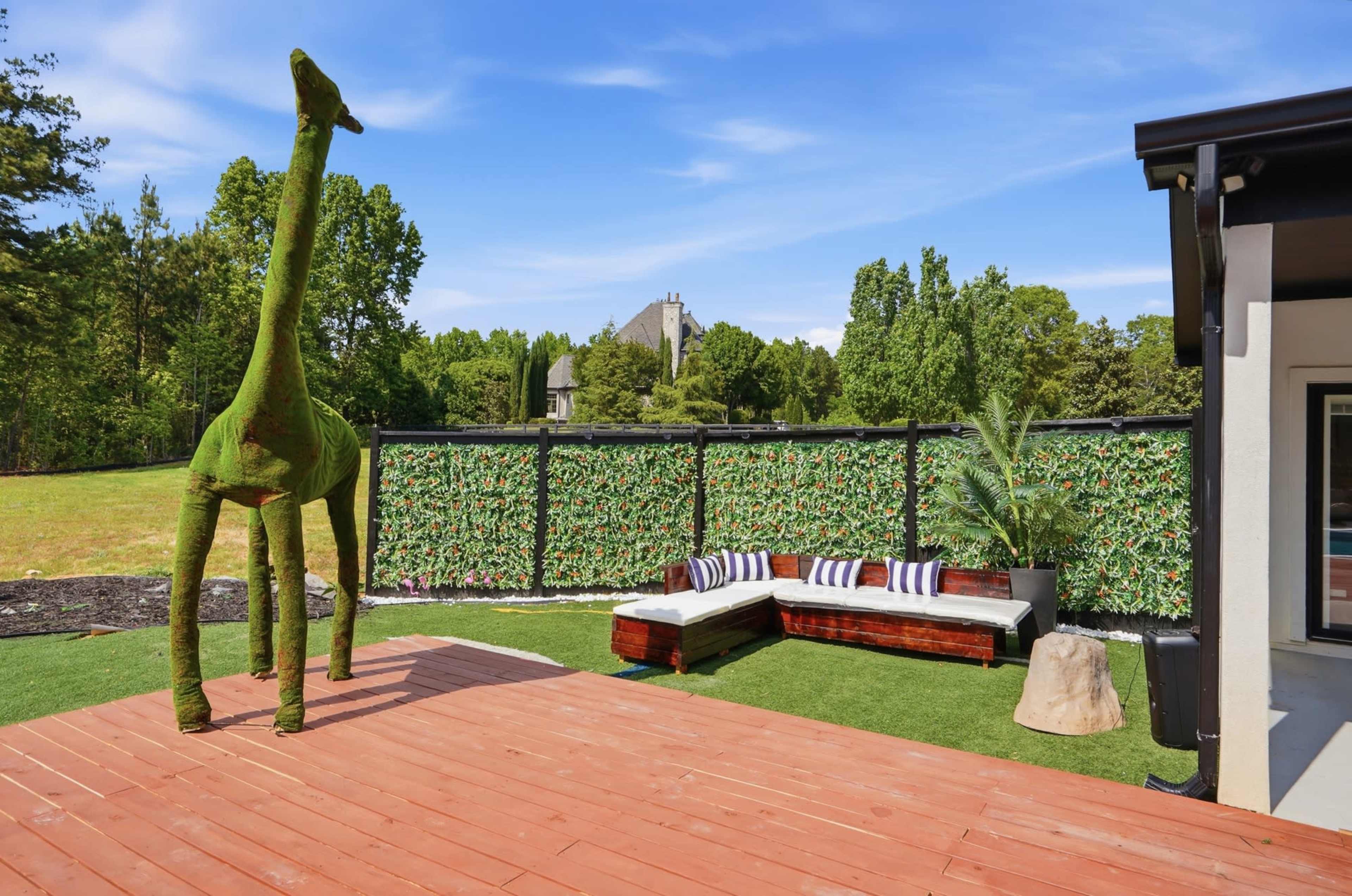 A modern outdoor living space features a green giraffe sculpture, a wooden deck, and a cushioned seating area surrounded by greenery.