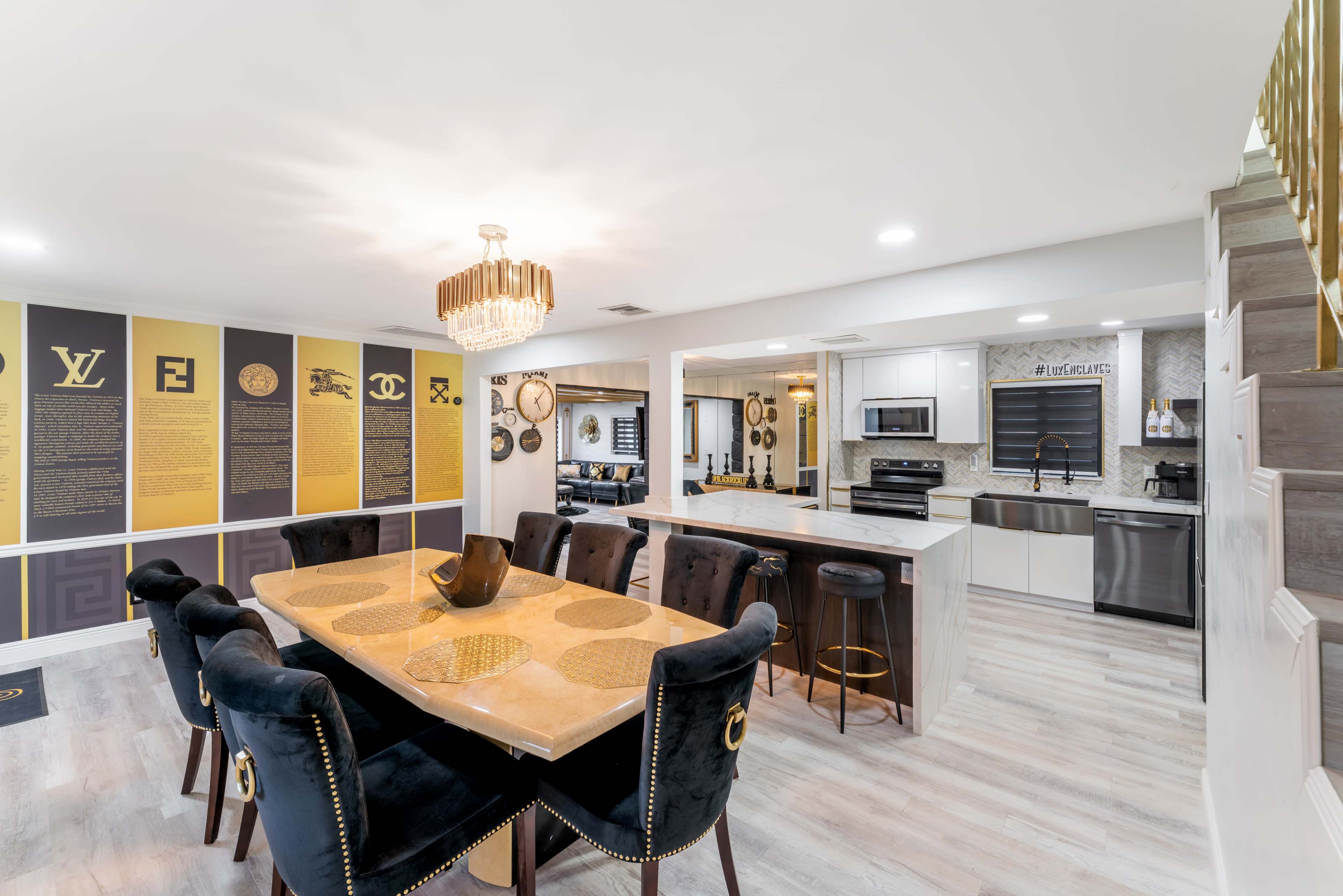 The image depicts a modern open-concept dining area featuring a large table with black upholstered chairs, a contemporary chandelier, and a kitchen with stainless steel appliances and a tiled backsplash.