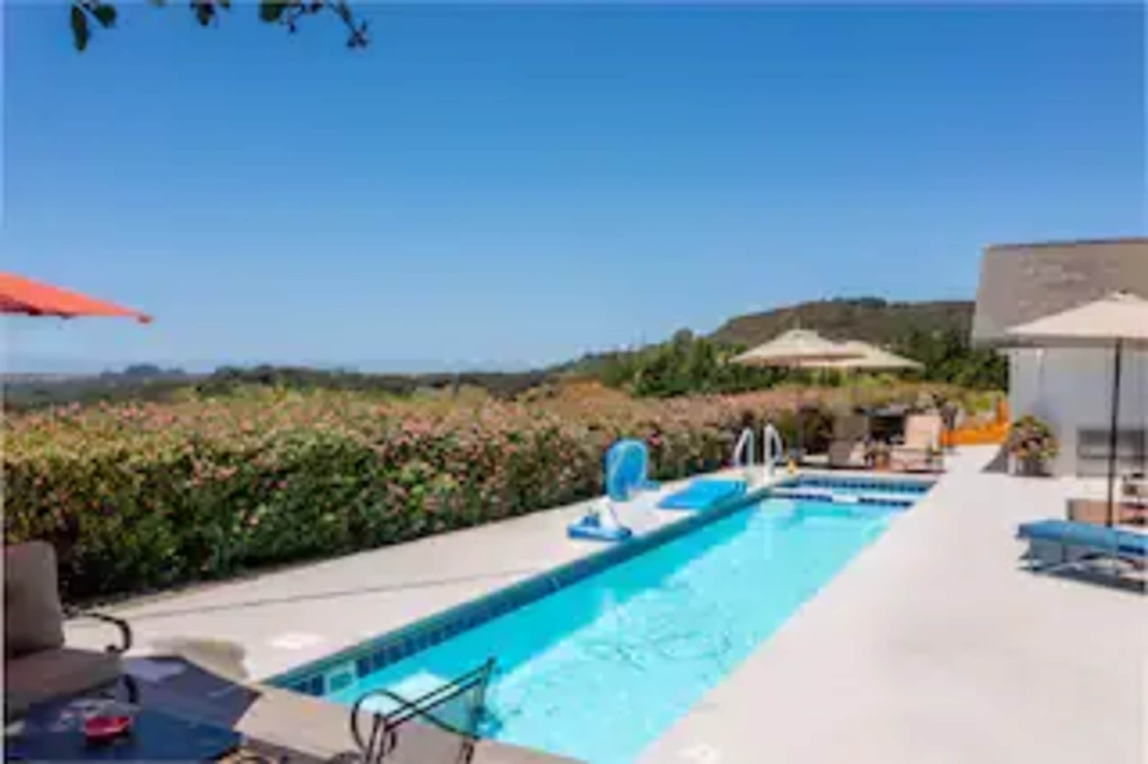 A swimming pool is surrounded by lounge chairs and umbrellas, with a backdrop of lush greenery and hills.