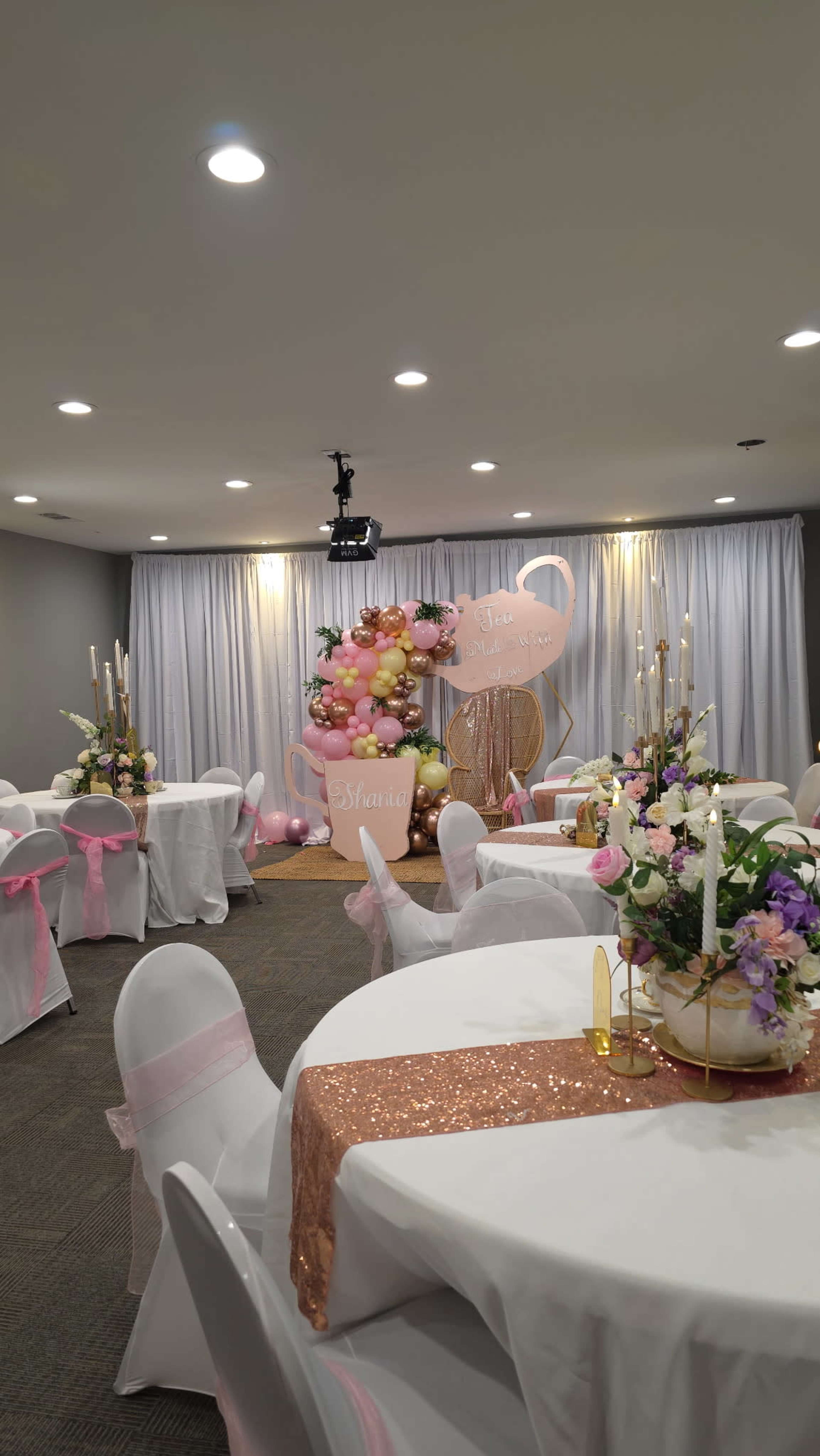 The image shows a decorated event space with round tables covered in white tablecloths and pink bows, adorned with floral centerpieces and a balloon arch against a backdrop.