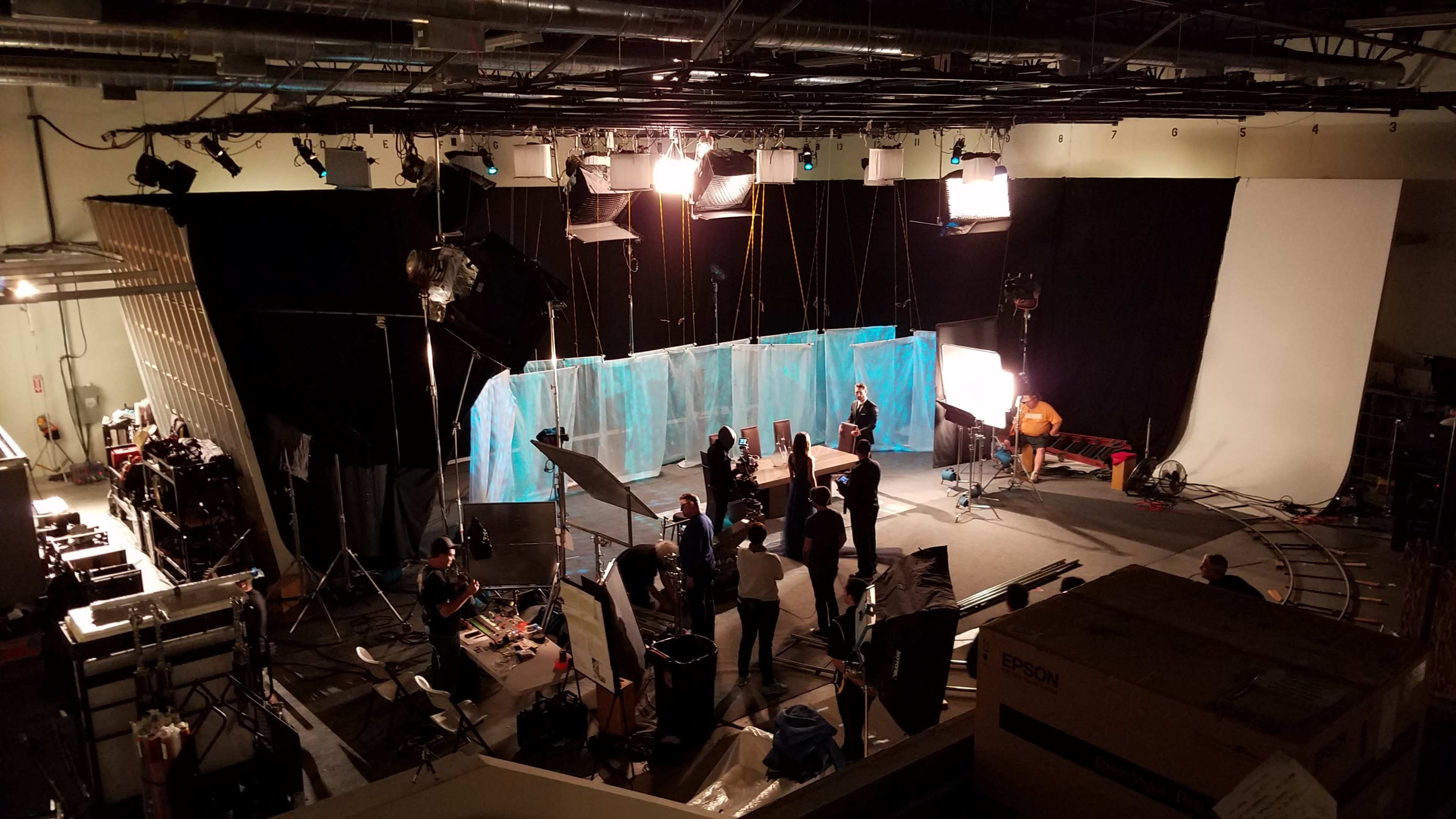 A film set is shown with crew members setting up equipment and lighting against a backdrop of blue and white screens.