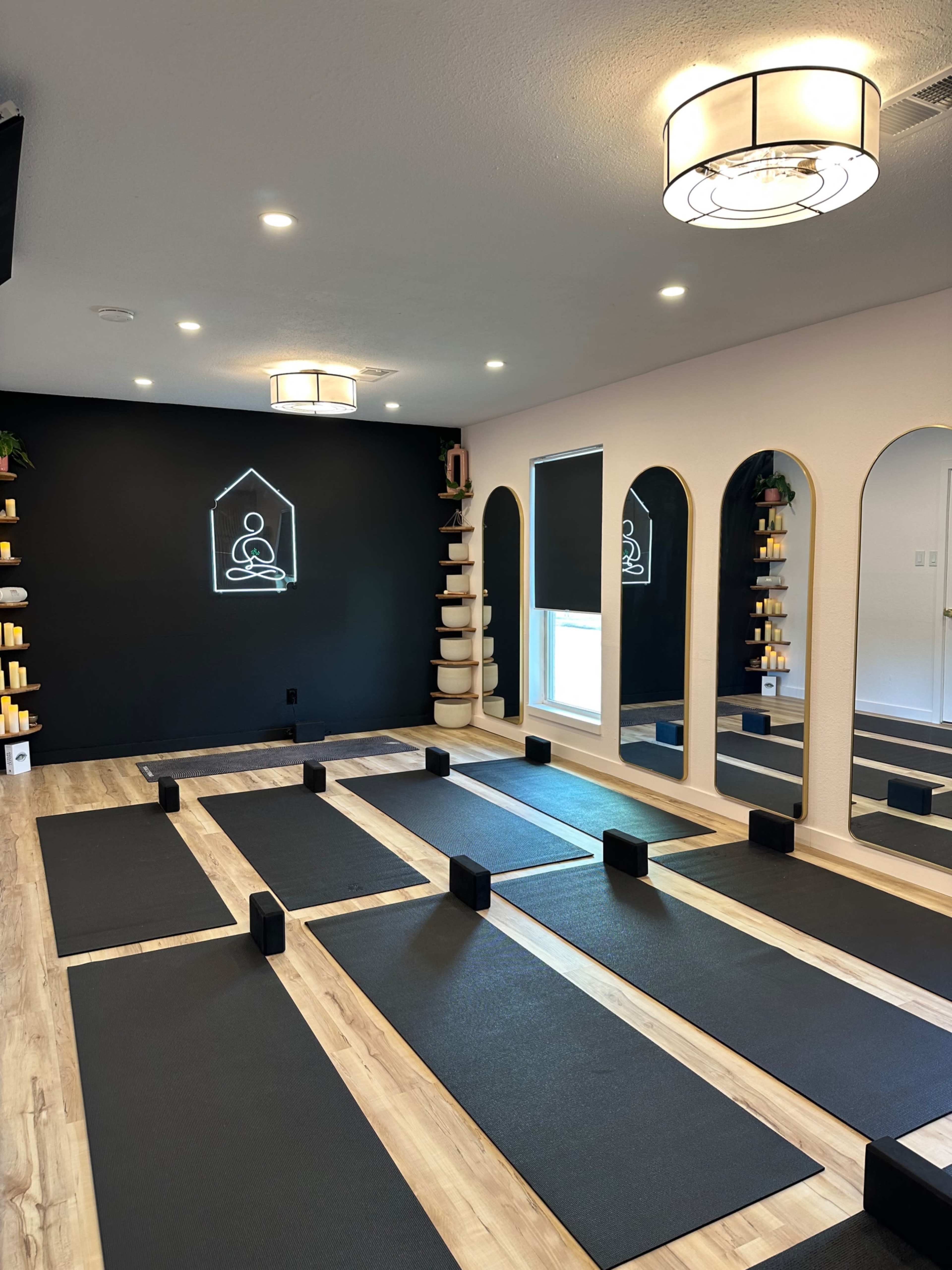 The image shows a minimalist yoga studio with black exercise mats arranged neatly on a wooden floor, mirrored walls, and a serene dark backdrop displaying a simple graphic of a seated figure.