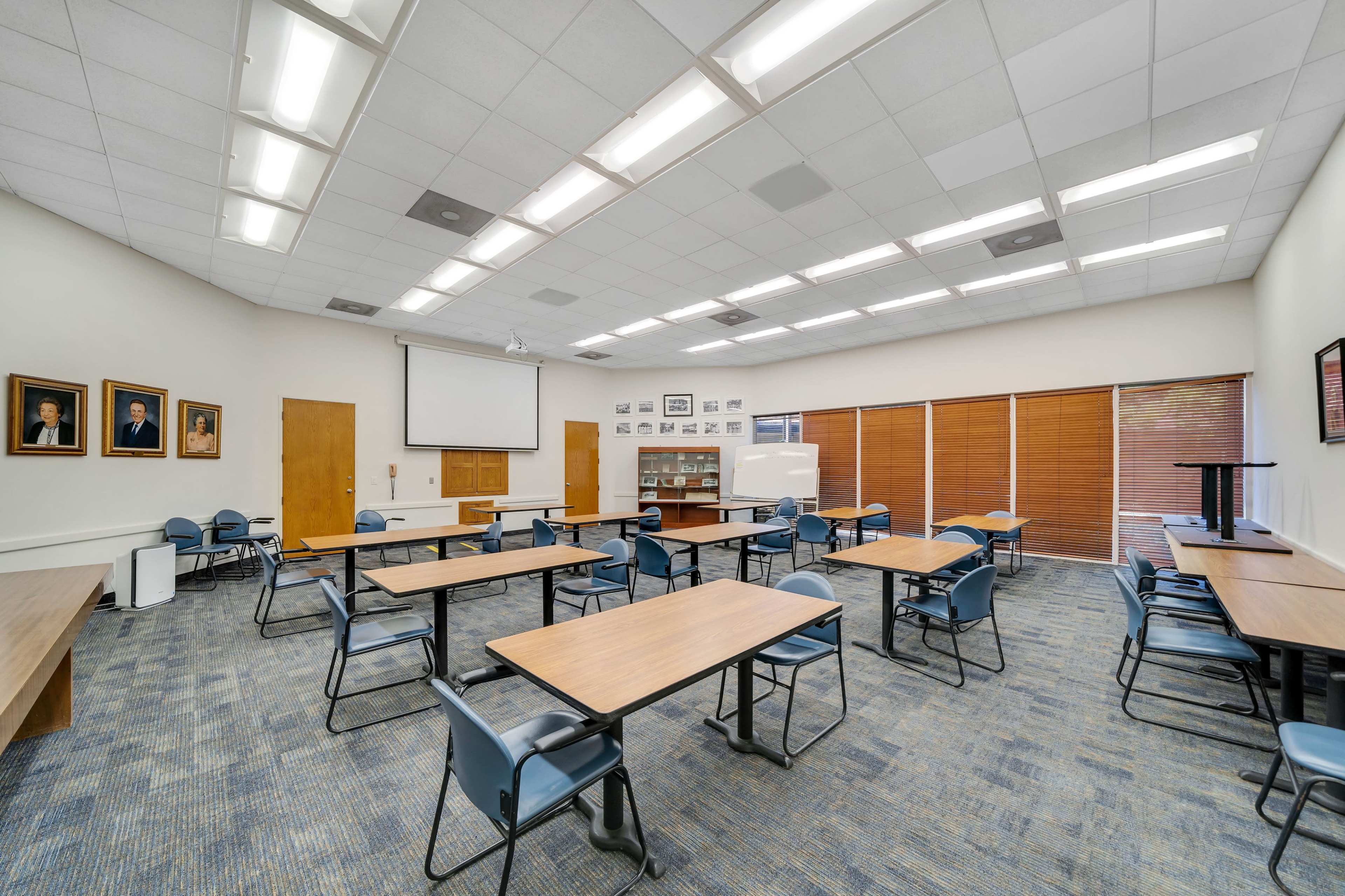 A classroom with several tables and chairs arranged, featuring a projector screen and multiple framed portraits on the walls.