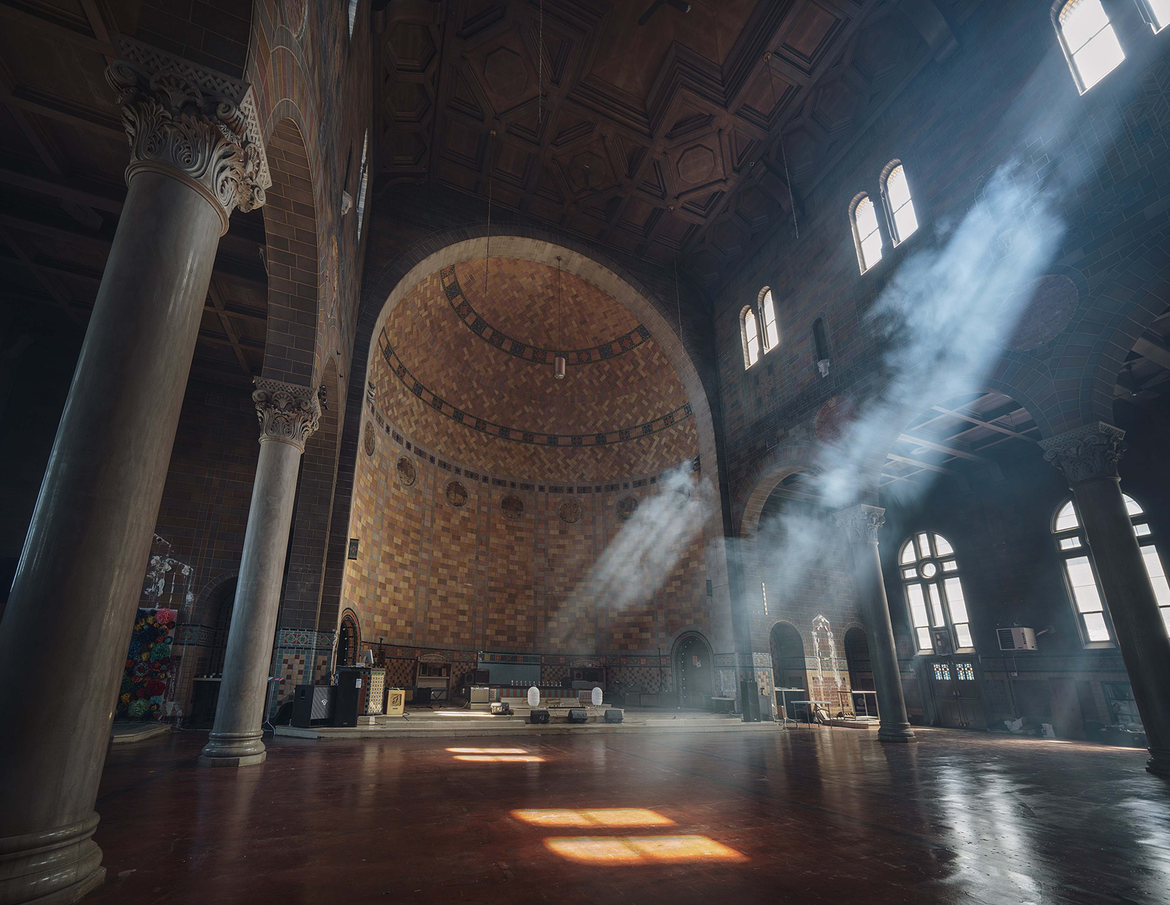 The interior of a grand, sunlit hall features high arches, ornate columns, and shafts of light illuminating the wooden floor.