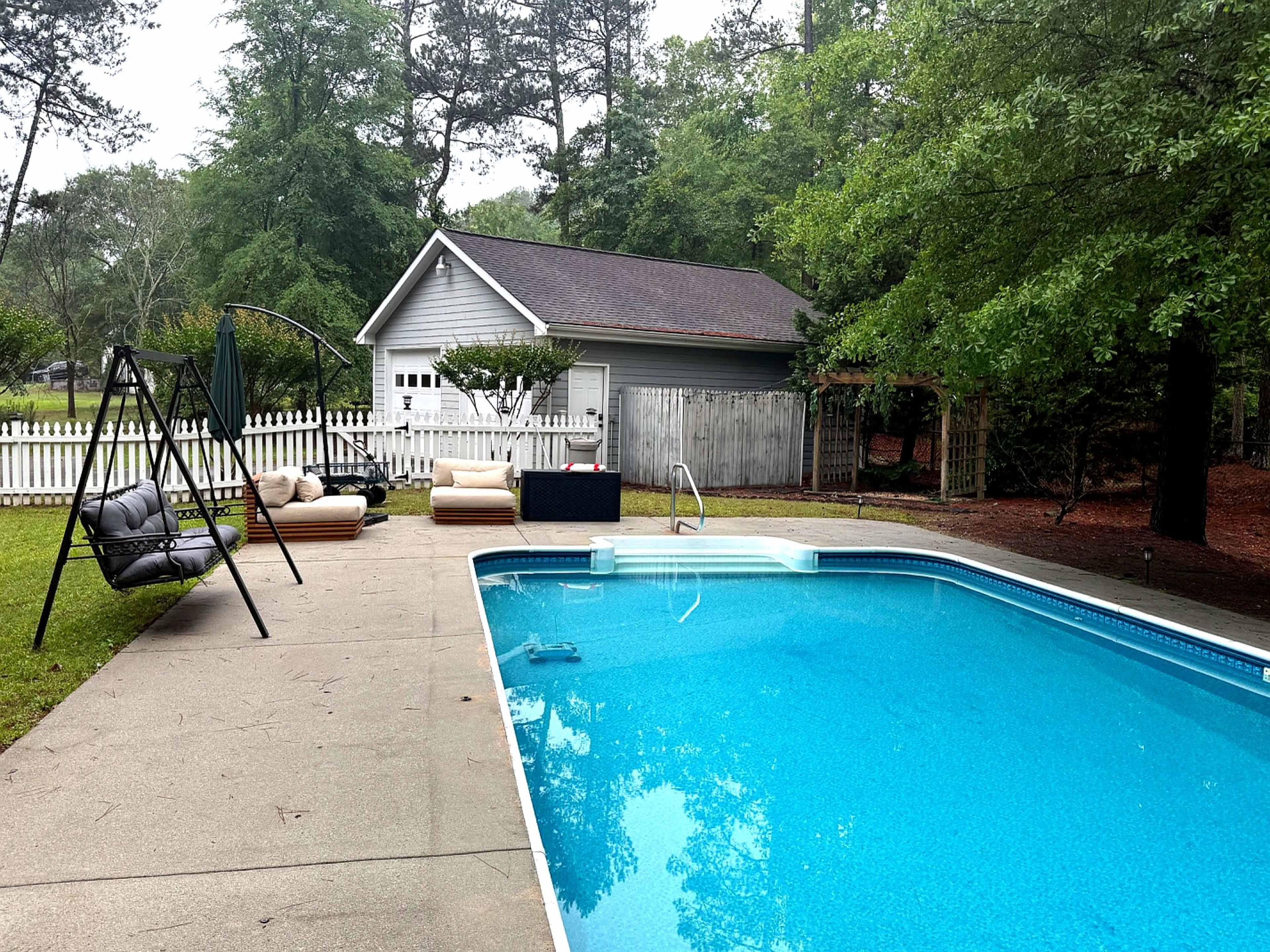 The scene features a swimming pool surrounded by a patio area with outdoor seating, a swing chair, and a shed in the background, all set within a wooded backyard.