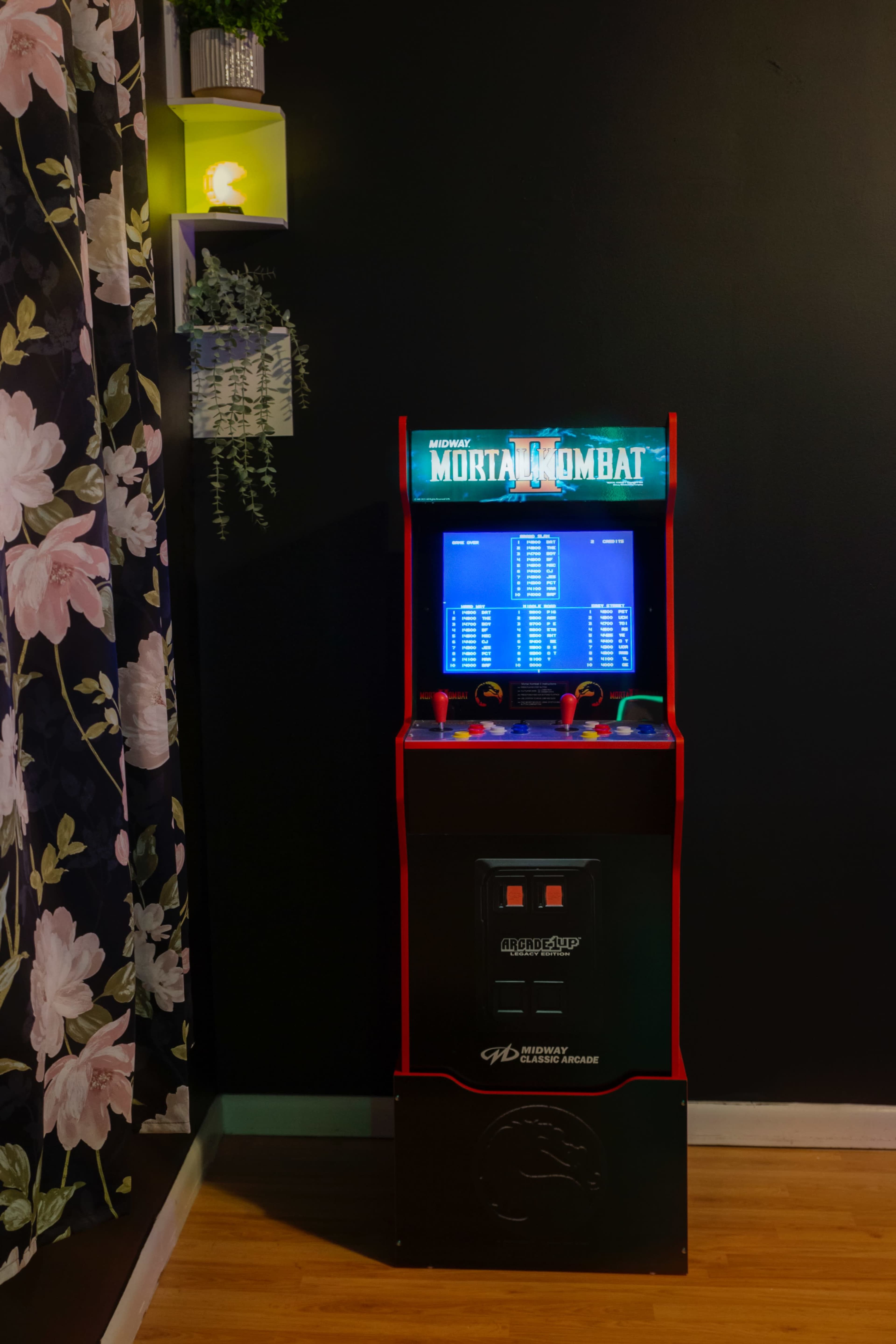 The image shows a retro Mortal Kombat arcade machine placed against a dark wall adorned with floral wallpaper.