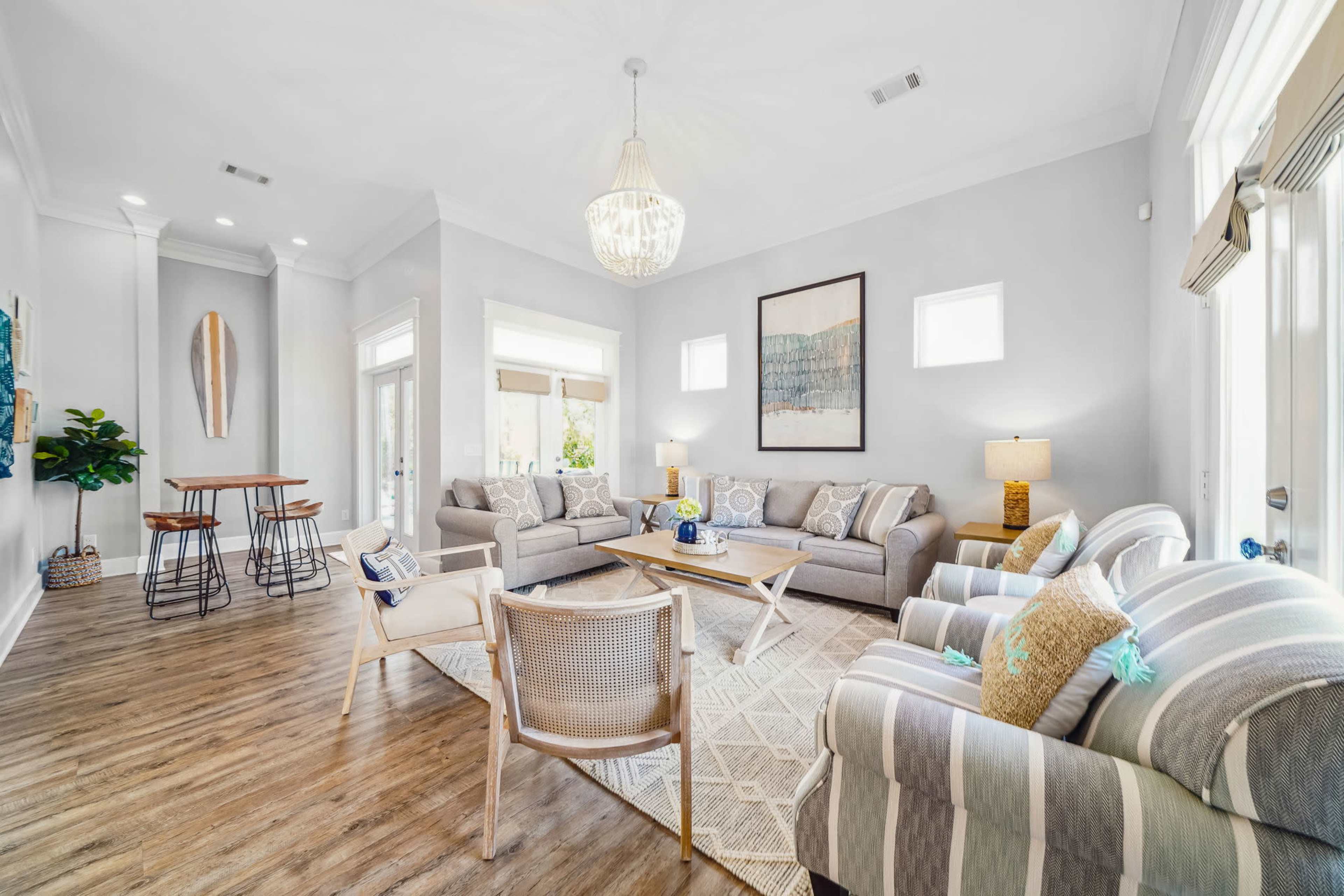 A bright and airy living room features gray sofas, a light wood coffee table, and a dining area with barstools, all set against a backdrop of large windows.