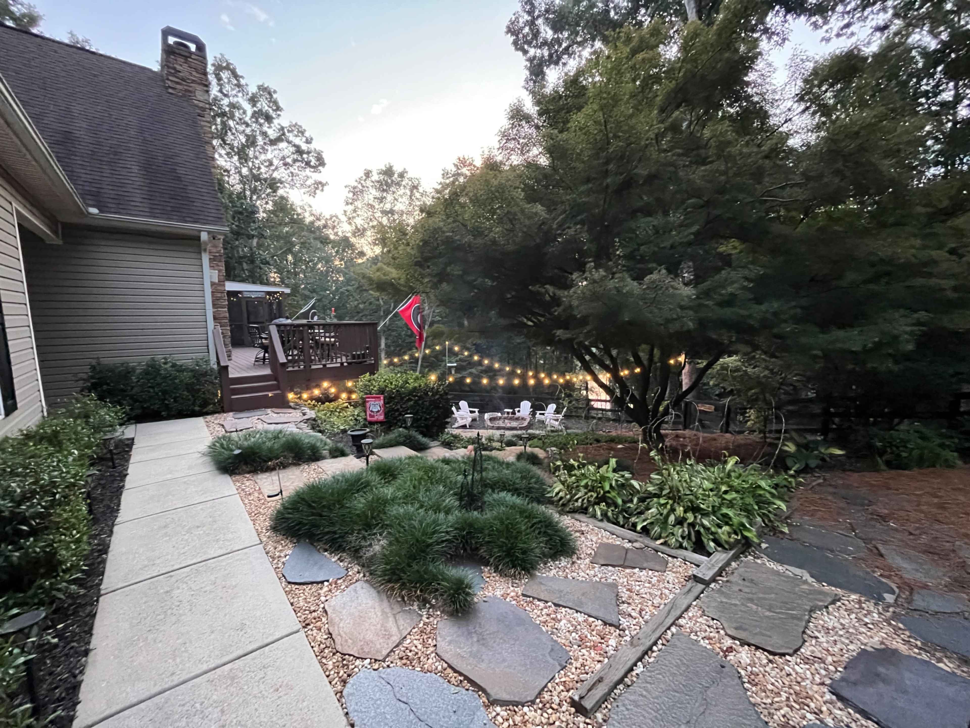 The image shows a pathway leading to a deck area surrounded by trees and landscaped garden features, including stone and plant arrangements.