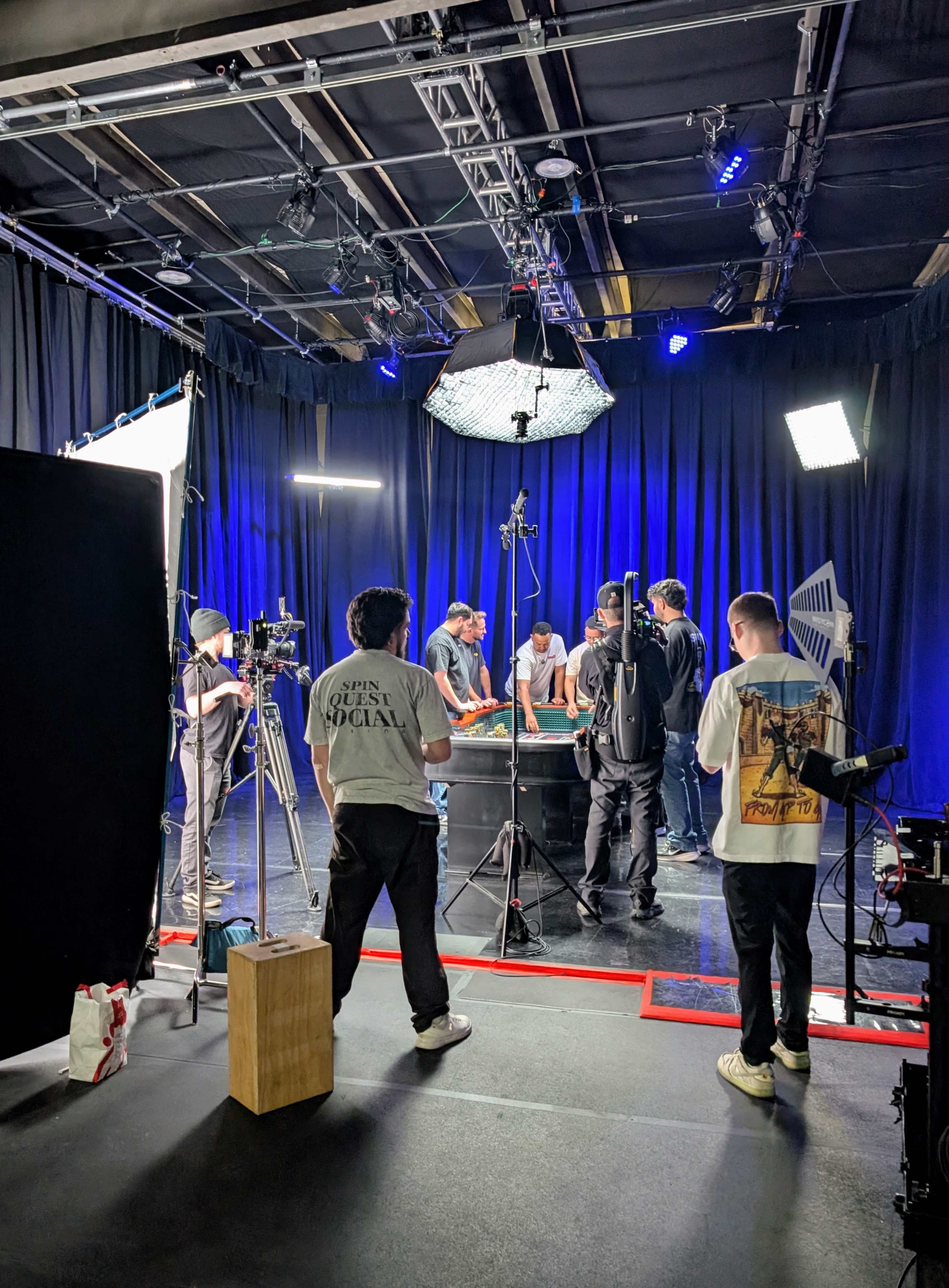 A group of people is gathered around a table in a studio, with various cameras and lighting equipment set up for a filming session.