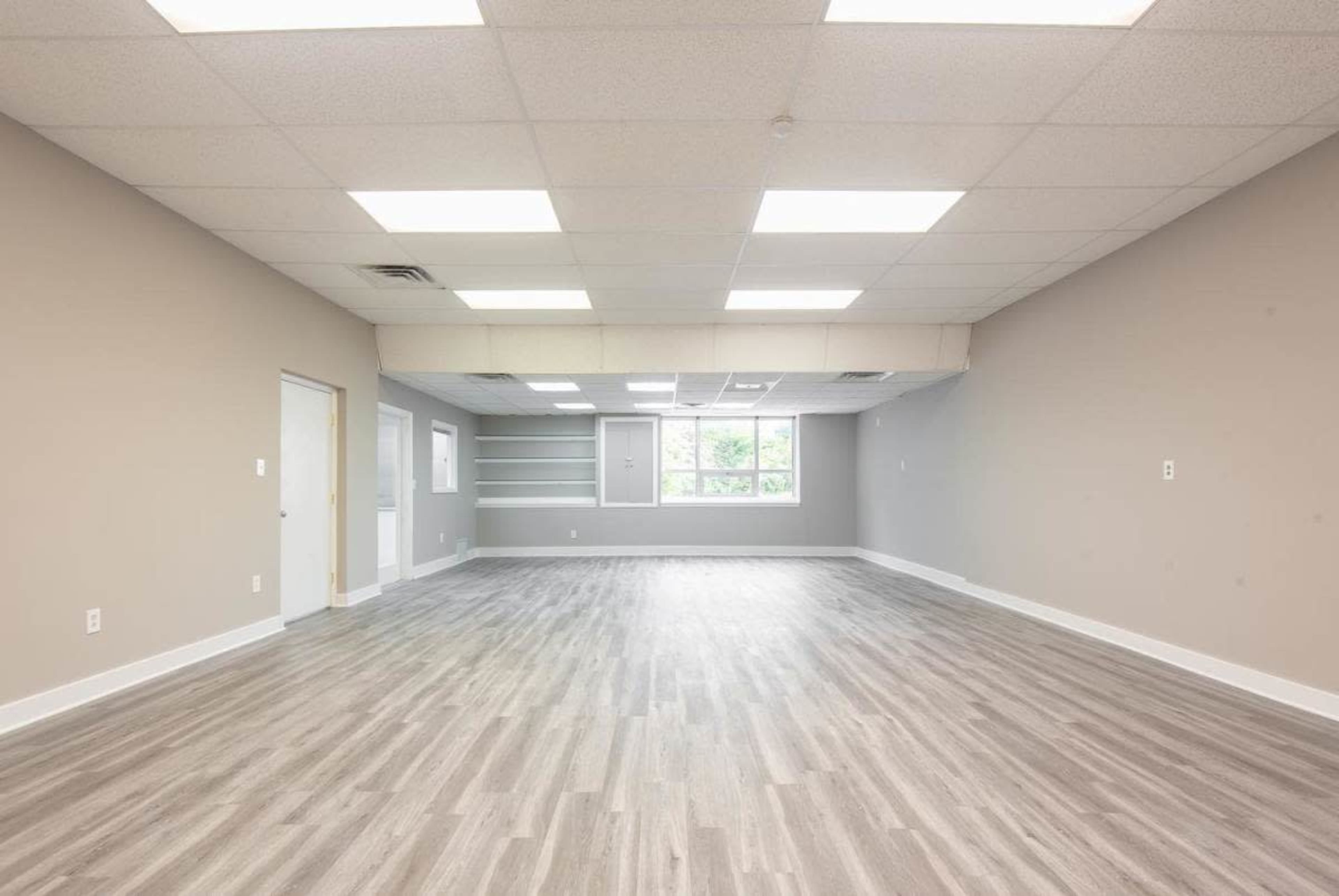 A spacious, empty room with gray walls and light-colored wooden flooring, featuring fluorescent ceiling lights and a window.
