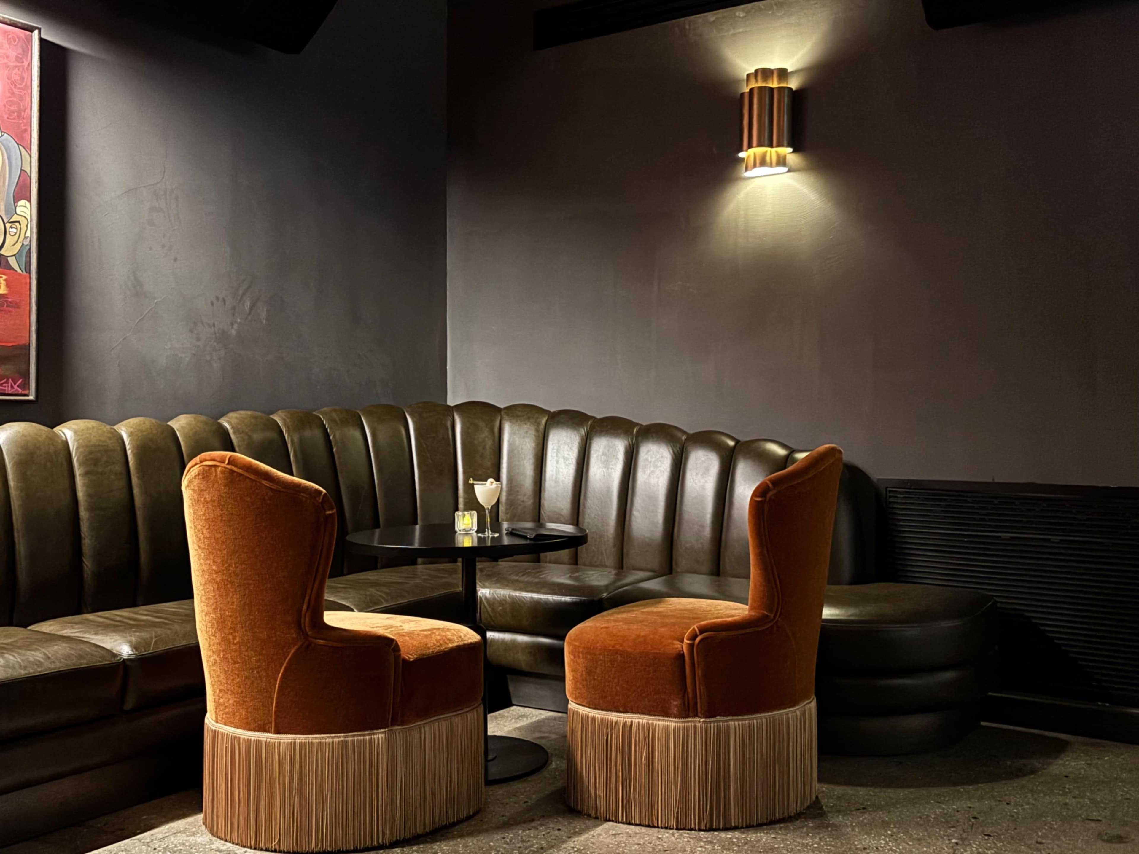 The image shows a cozy seating area with a curved brown leather sofa, a round black table, and two orange chairs with fringe details.