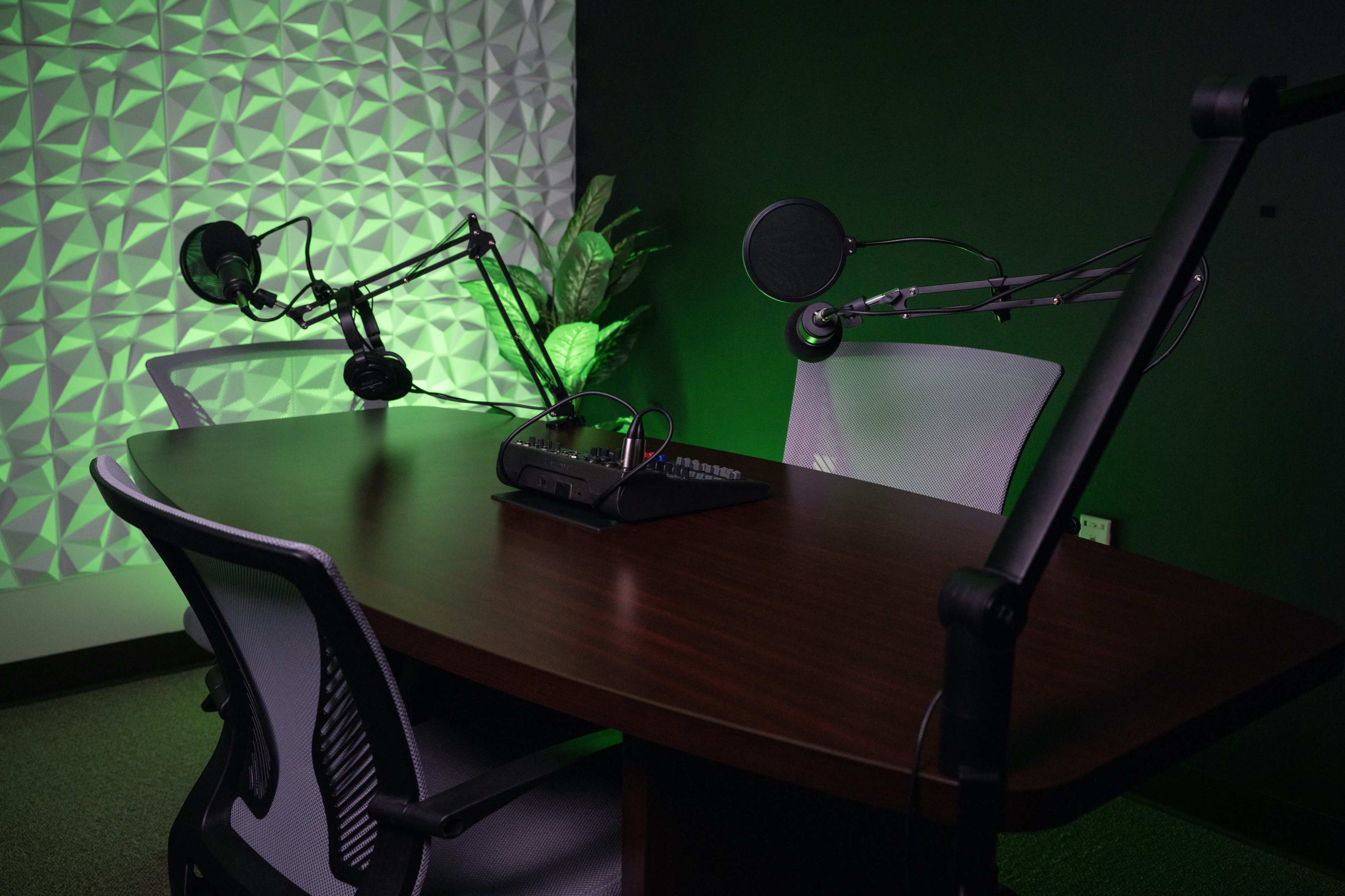 A podcast recording studio featuring a wooden table, two microphone setups on adjustable arms, and green ambient lighting.
