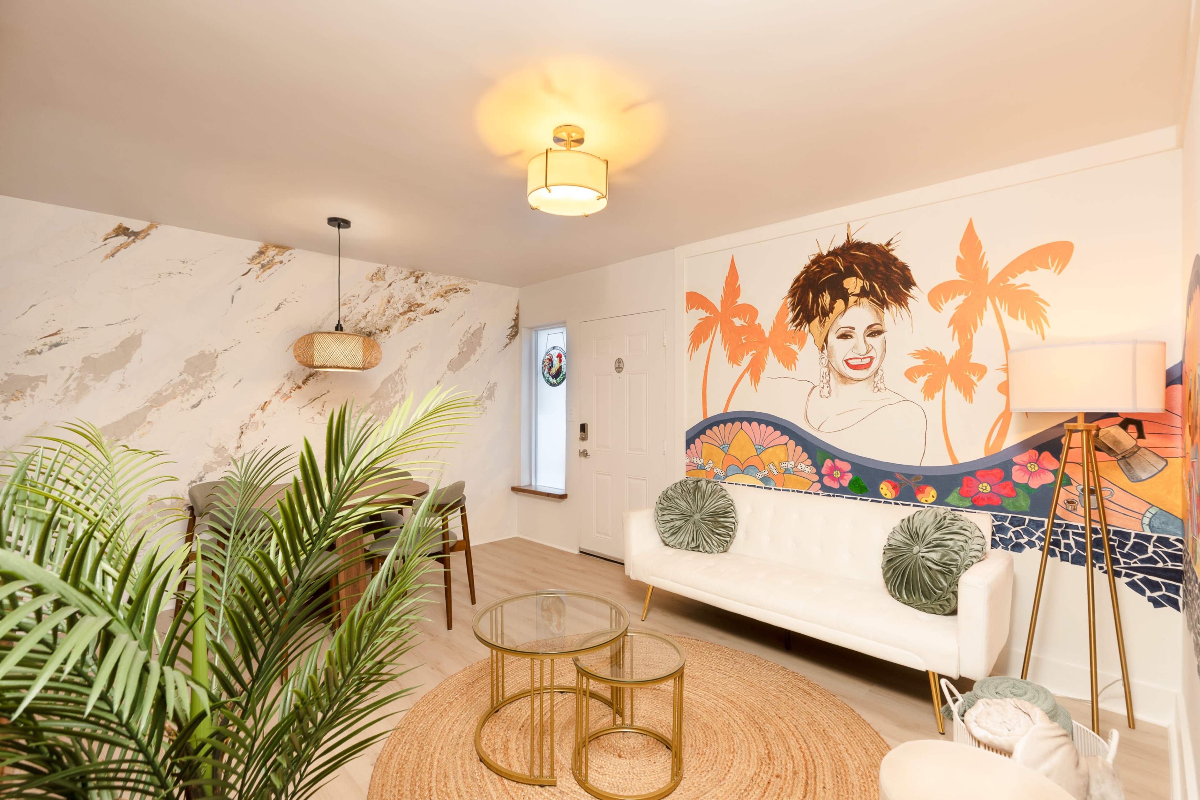 The image shows a modern living room featuring a white sofa, a round area rug, a coffee table, and a mural with a woman and tropical plants on one wall.