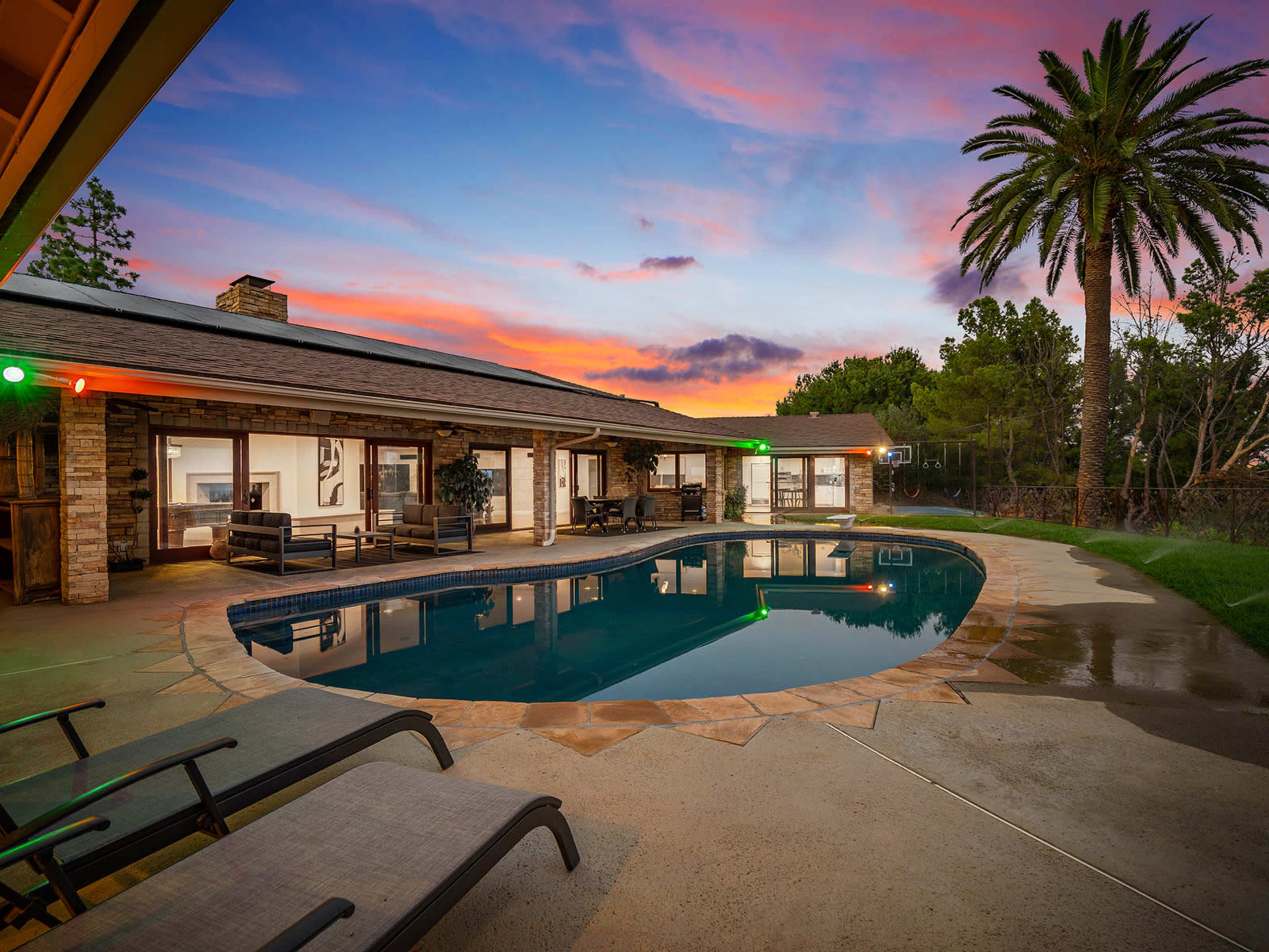 A modern home with a swimming pool reflects a vibrant sunset, surrounded by palm trees and outdoor seating.