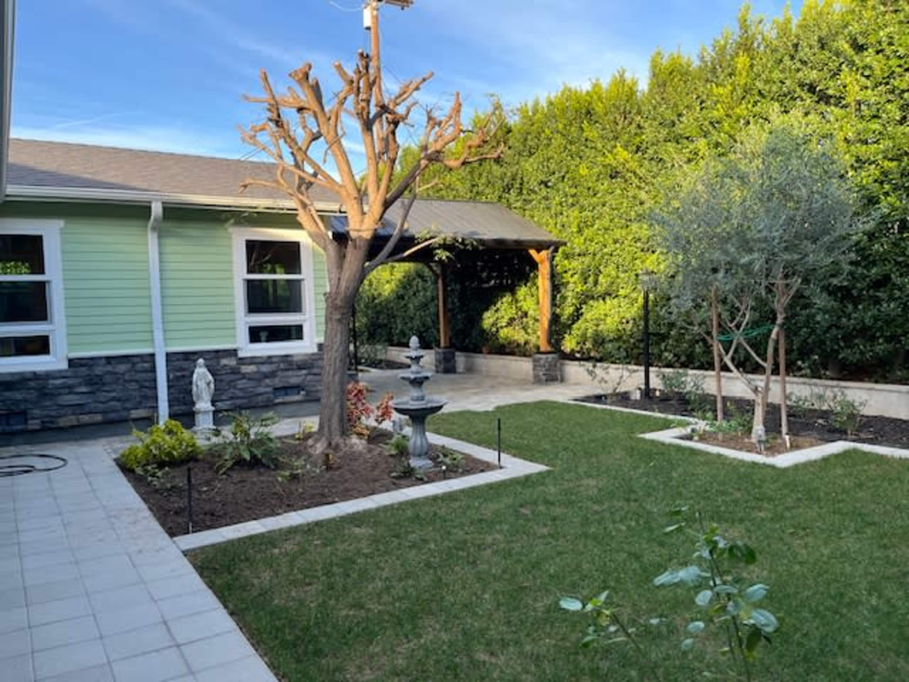 The image features a landscaped backyard with a stone path, a central ornamental tree, garden beds, a gazebo, and a statue amidst greenery.