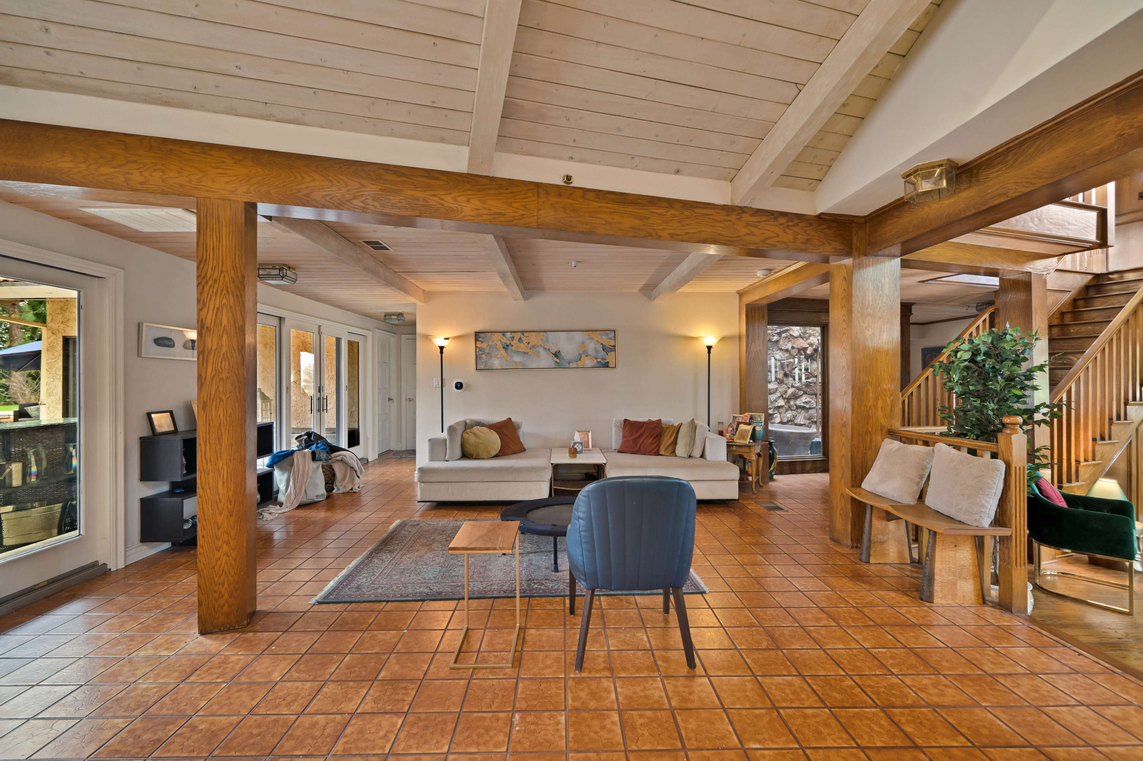 The image shows a spacious living room with wooden beams, a tiled floor, and a staircase leading to an upper level.