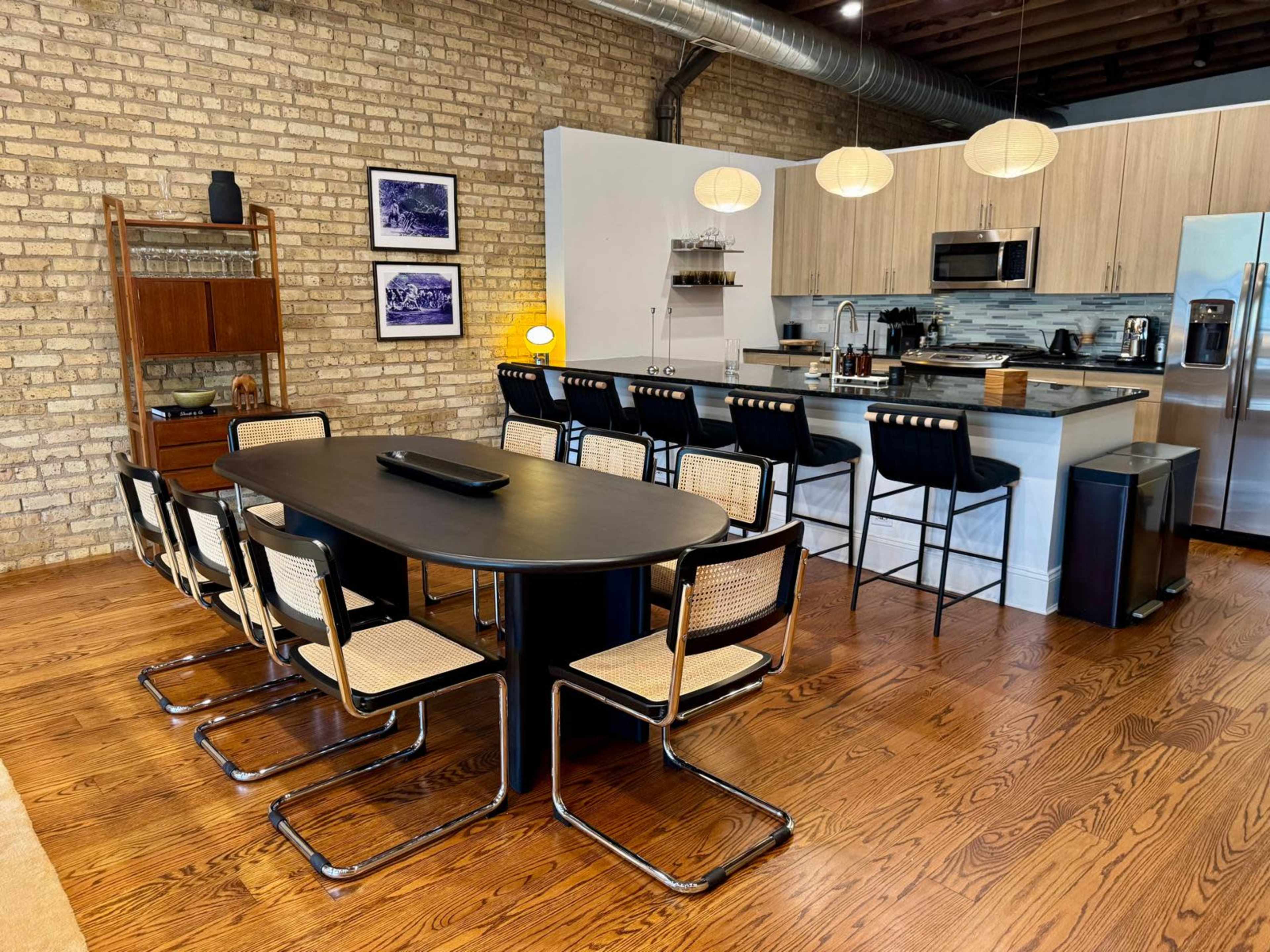 The image shows a modern kitchen and dining area with a round black table surrounded by chairs, and a kitchen island with bar stools, all set in a spacious room with exposed brick walls.