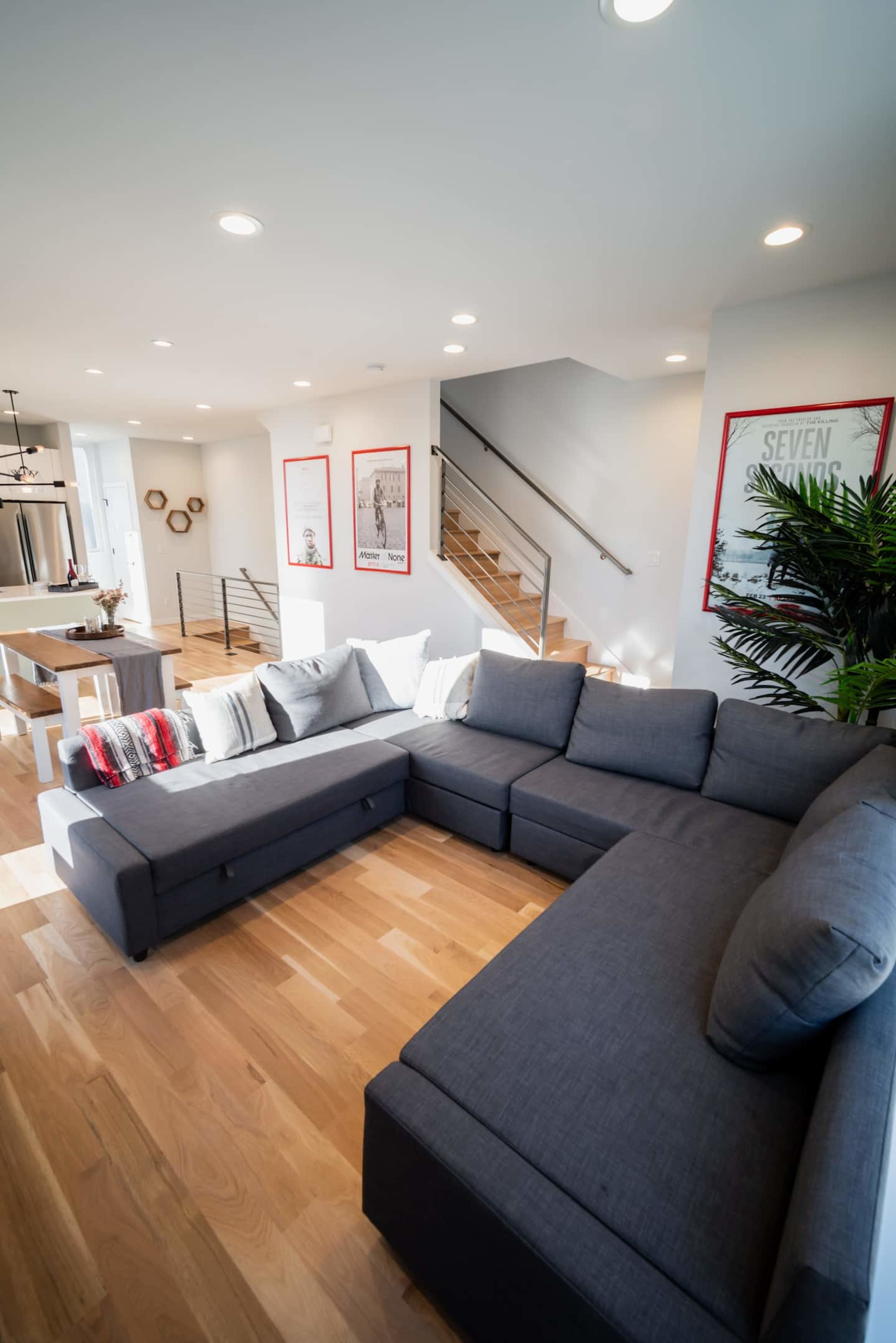 A gray sectional sofa is positioned in a bright living room with hardwood flooring, surrounded by framed wall art and a staircase in the background.
